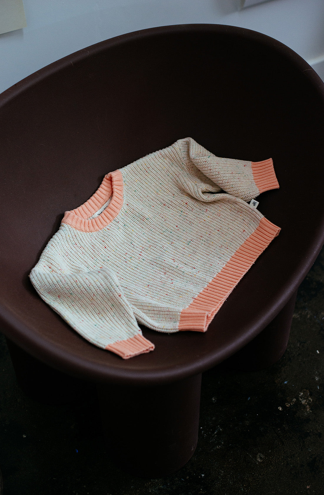 A Freckle Knit Peach jumper from SUNDAY SIBLINGS is laid out on a dark brown armchair. The 100% cotton sweater, featuring a stylish beige and pink design with a pink ribbed collar, cuffs, and hem for a comfortable fit, displays a simple pattern and has a tag on the side. The chair rests on a dark floor in an illuminated room.