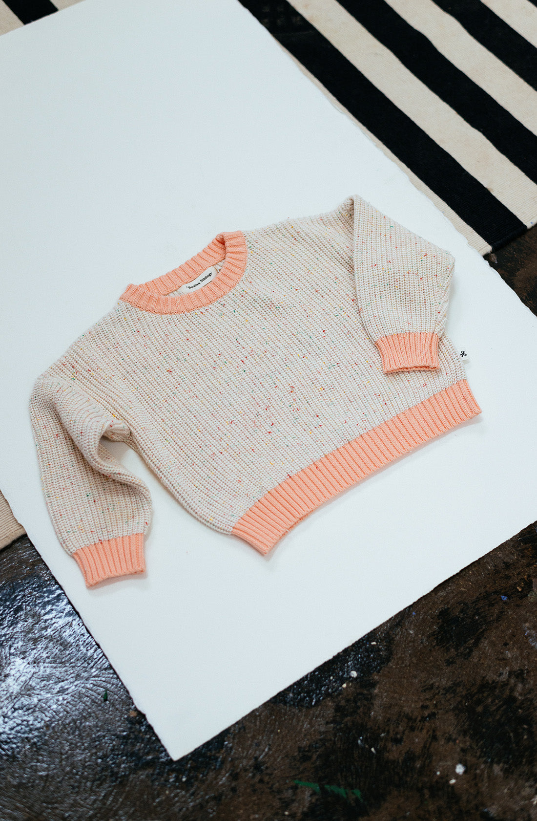 A cream-colored Freckle Knit Peach jumper from SUNDAY SIBLINGS, featuring colorful speckles and peach-colored ribbed cuffs, hem, and collar, lies flat on a white and black striped surface. Crafted from 100% cotton, it offers a comfortable fit that's perfect for any occasion.