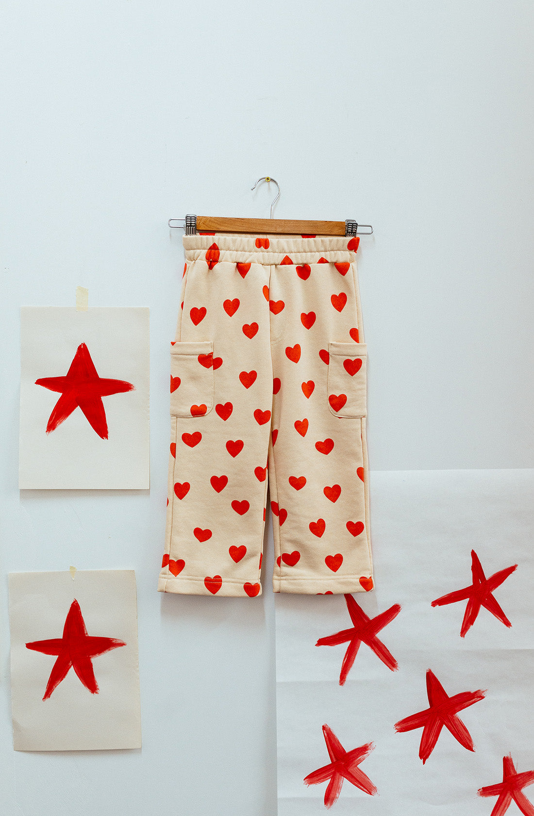 SUNDAY SIBLINGS' Holiday Pants Red Hearts, made from luxurious French terry cotton, hang on a clothes hanger against a white wall. These ultra-soft beige sweatpants with red heart patterns are framed by three papers taped to the wall: one featuring a single red star, another adorned with multiple red stars, and a third partially off-screen paper displaying one star.