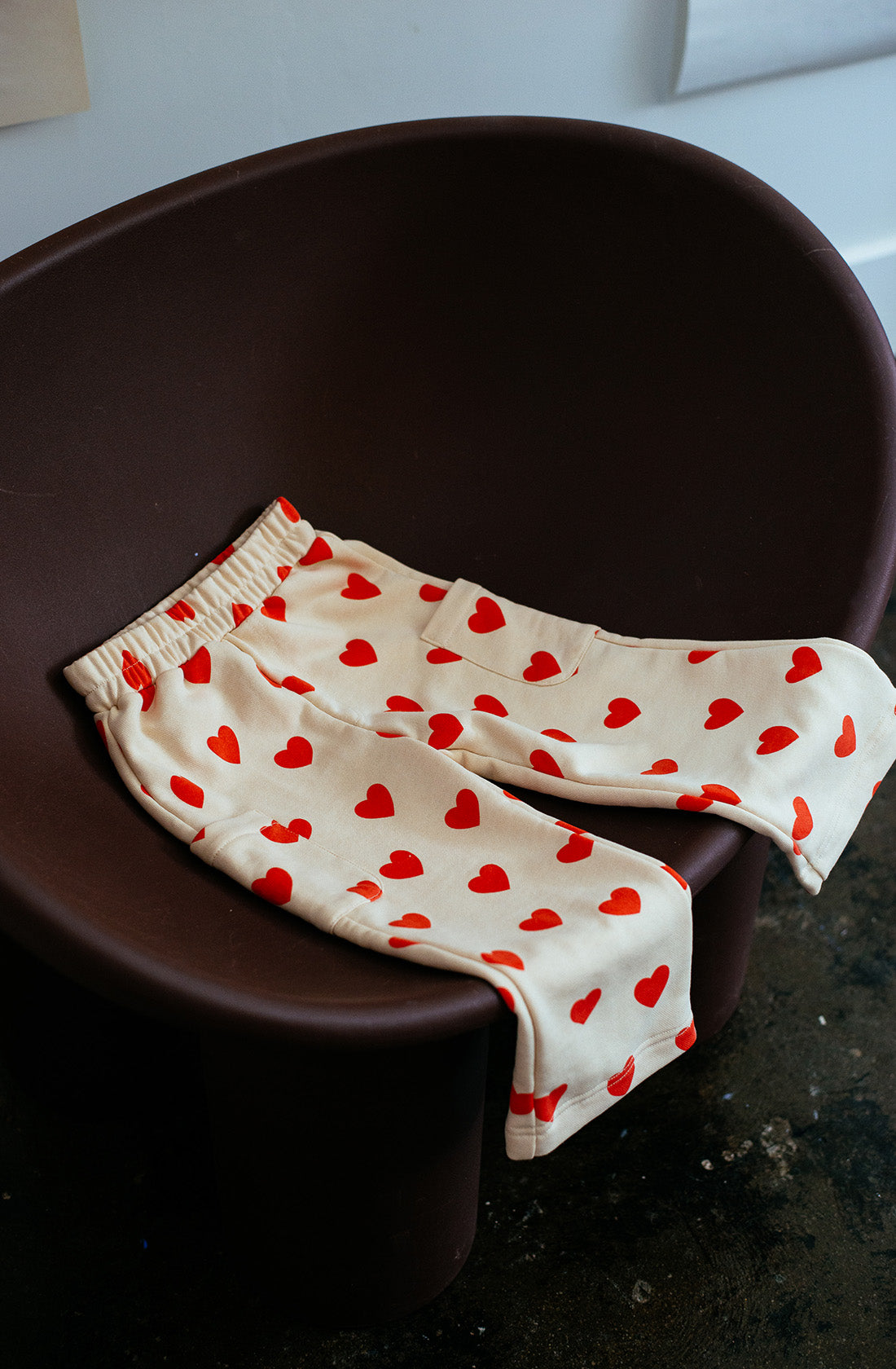 A pair of SUNDAY SIBLINGS' Holiday Pants Red Hearts, made from ultra-soft French terry cotton and featuring a beige color with red heart patterns, is draped over a dark brown, modern chair in an indoor setting with a visible dark floor.