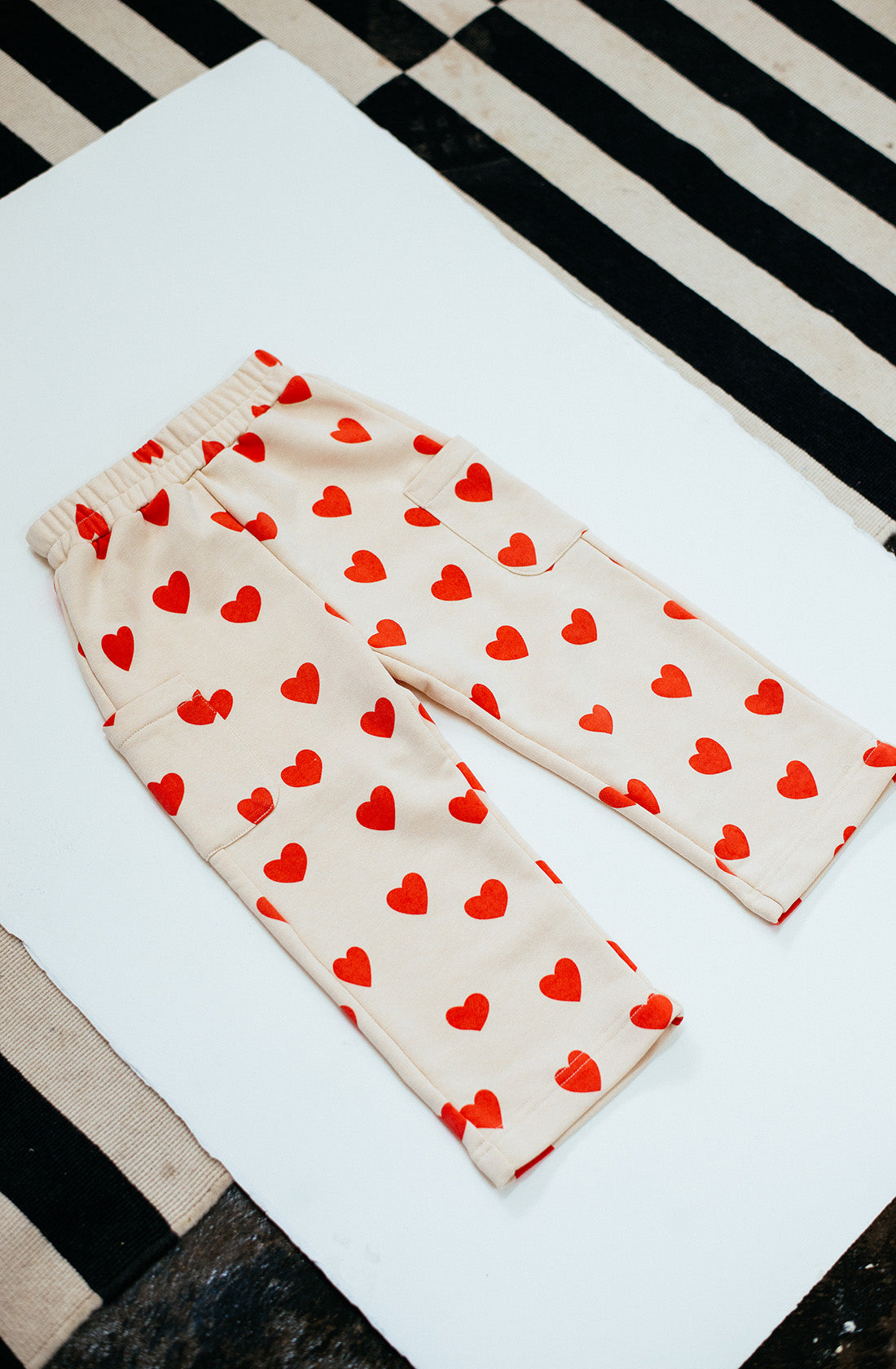 A pair of Holiday Pants Red Hearts by SUNDAY SIBLINGS, featuring an elastic waistband and adorned with a pattern of red hearts on beige, is laid flat on a white surface. Made from ultra-soft French terry cotton fabric that promises comfort, the background showcases black and white striped textiles.