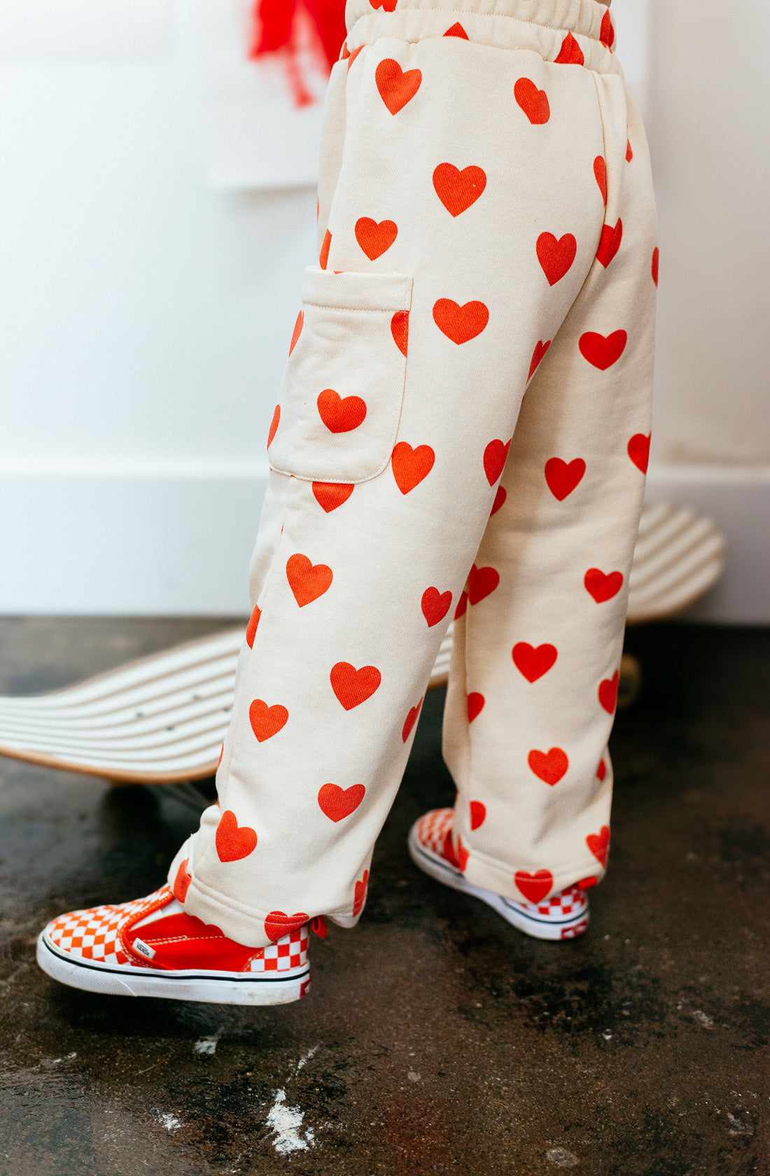 A child clad in SUNDAY SIBLINGS Holiday Pants Red Hearts and red and white checkerboard slip-on shoes stands on a skateboard. The scene appears to be indoors, with part of a white wall and floor visible in the background.