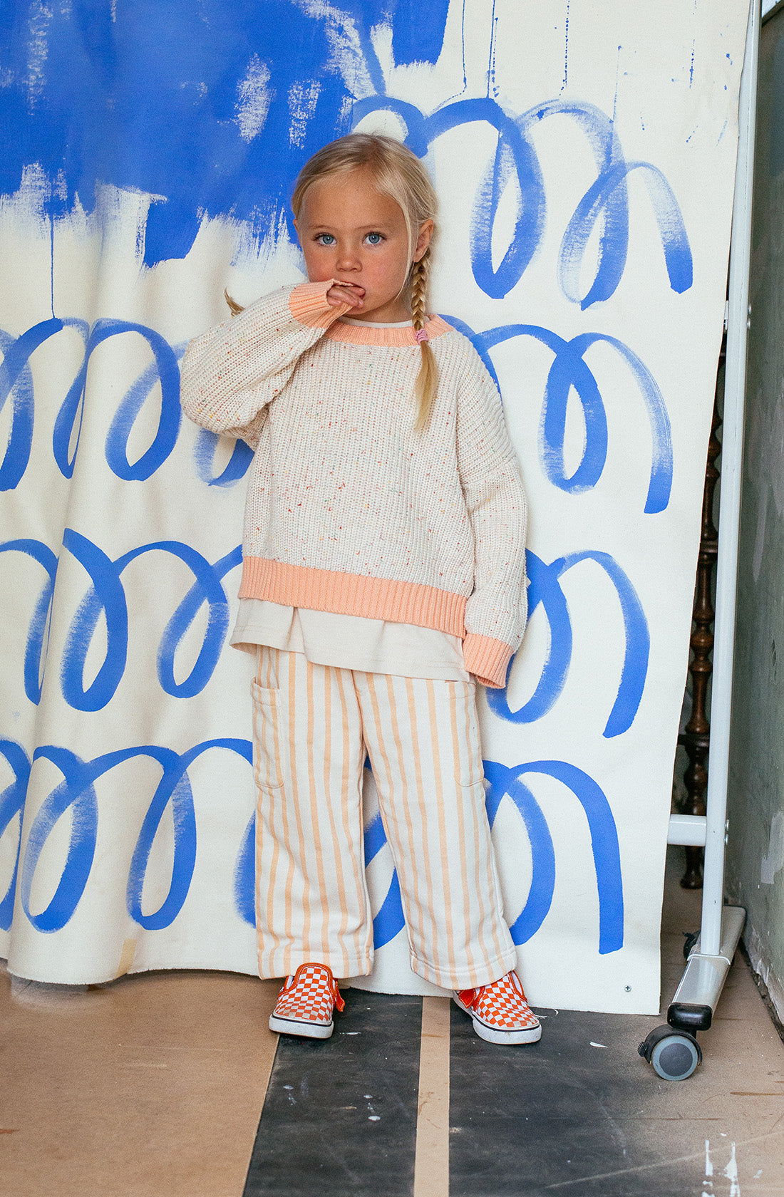 A young child with long, blonde hair in two braids stands in front of a white backdrop adorned with blue spiral patterns. They are wearing a cream Freckle Knit Peach jumper from SUNDAY SIBLINGS, paired with striped pants and orange-checkered shoes. The comfortable fit ensures they feel at ease, one hand near their mouth.