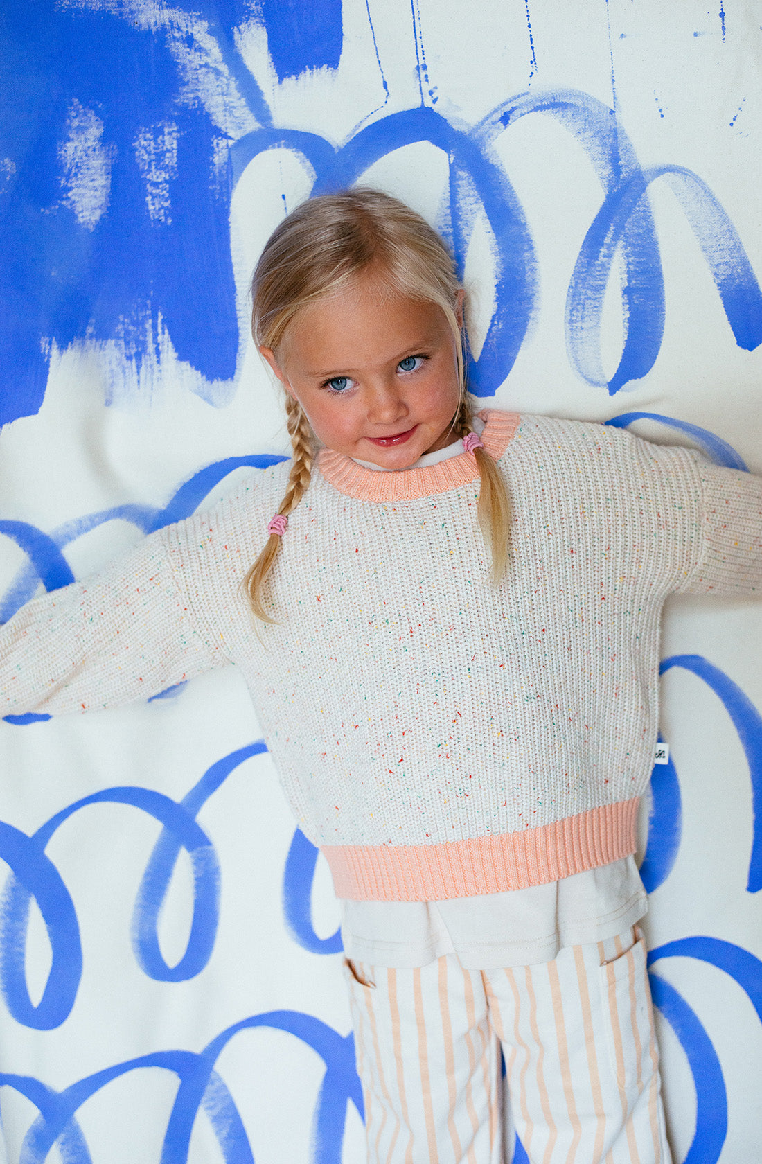 A young girl with blonde hair in two braids is wearing the Freckle Knit Peach jumper by SUNDAY SIBLINGS and striped pants. The comfortable fit of her 100% cotton outfit complements the playful expression on her face as she stands against a background painted with blue swirls and brush strokes, looking slightly off-camera.