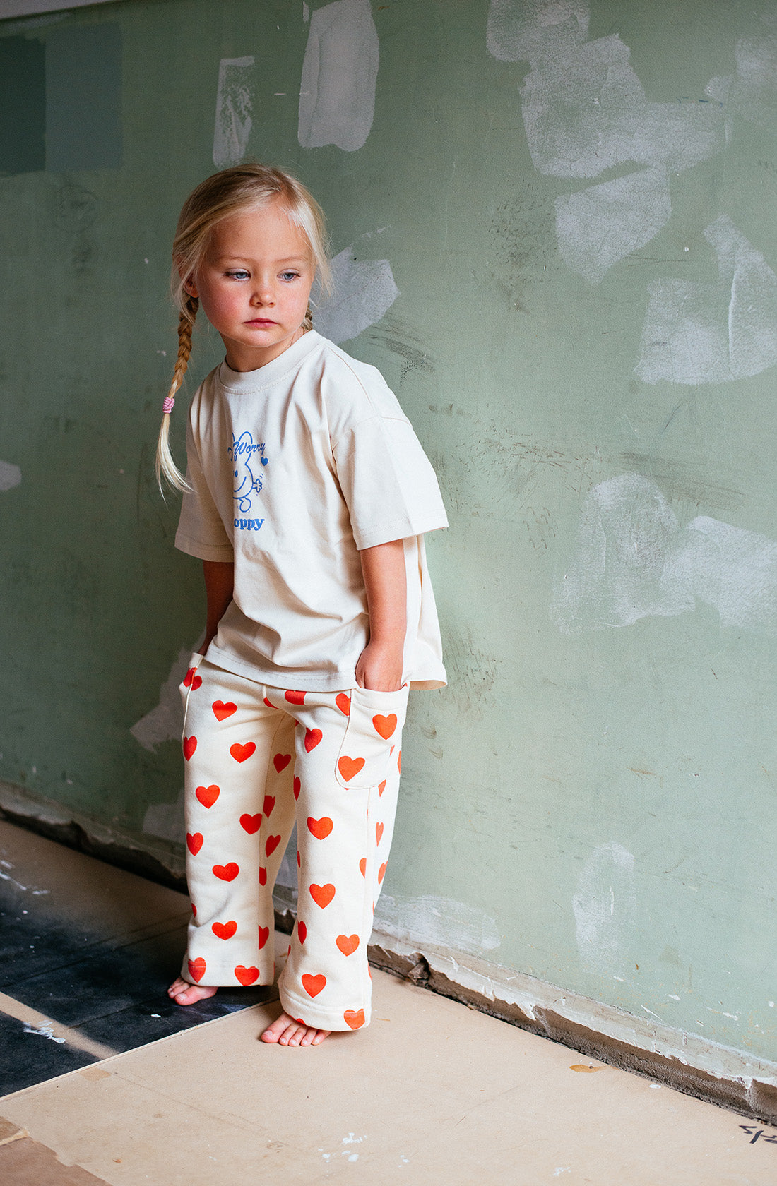 A young girl with blonde hair in a side braid stands barefoot against an unfinished wall, wearing an oversized beige T-shirt and ultra-soft Holiday Pants Red Hearts by SUNDAY SIBLINGS. The cream-colored pants, adorned with a red heart pattern, are made from French terry cotton. With her hands in her pockets, she wears a pensive expression.