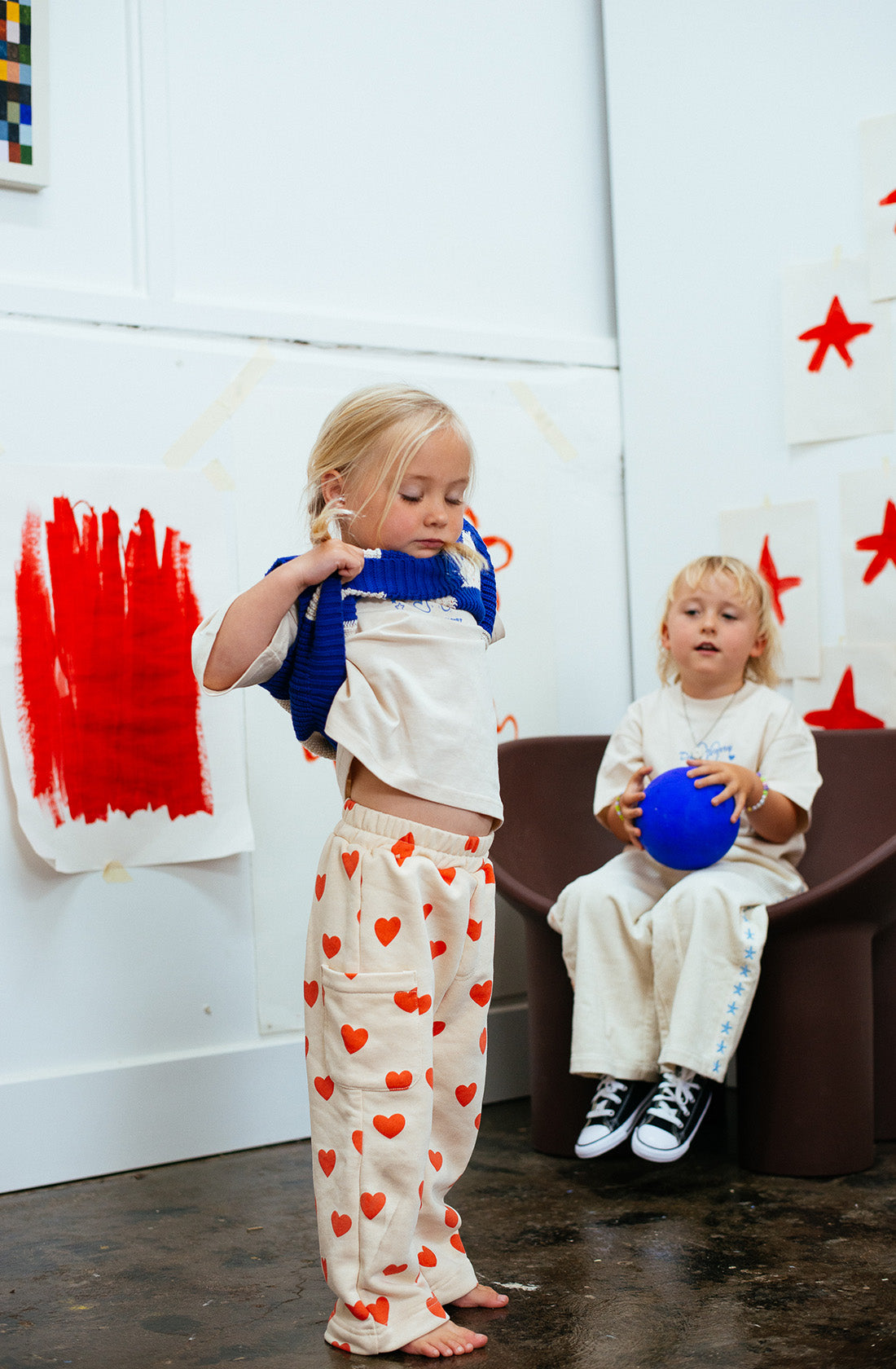 Two young children are in a playroom with artistic decor. One child is standing and putting on a blue sweater over a white shirt and SUNDAY SIBLINGS' Holiday Pants Red Hearts. The other child is sitting on a chair, holding a blue ball, with paint-covered paper on the wall behind them.