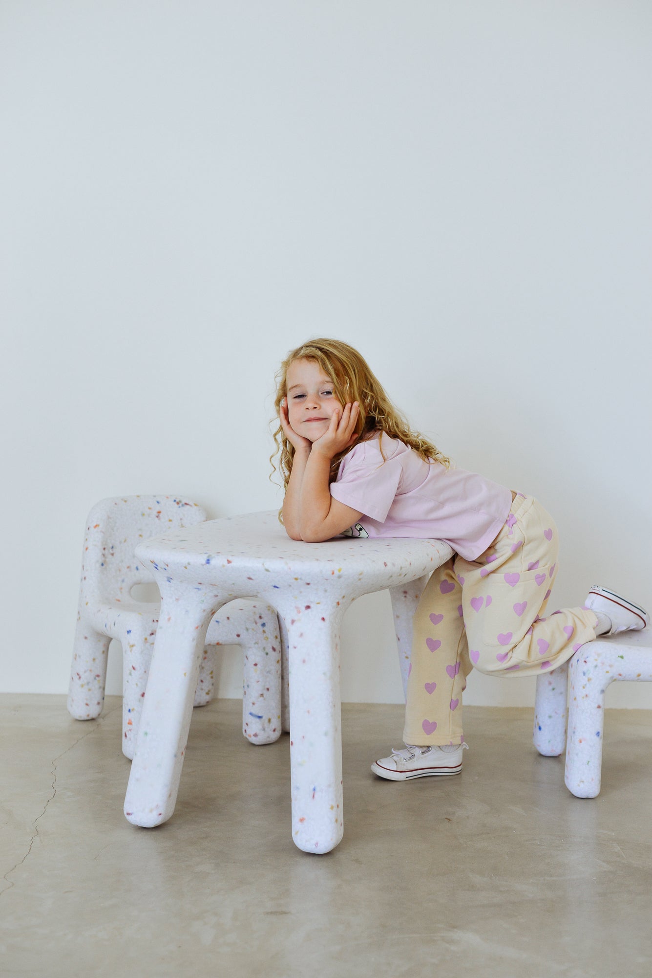 A young girl with long curly hair leans on a white speckled table wearing a light pink shirt, sneakers, and SUNDAY SIBLINGS Sunday Siblings - Holiday Pants Purple Hearts, gazing thoughtfully at the camera.