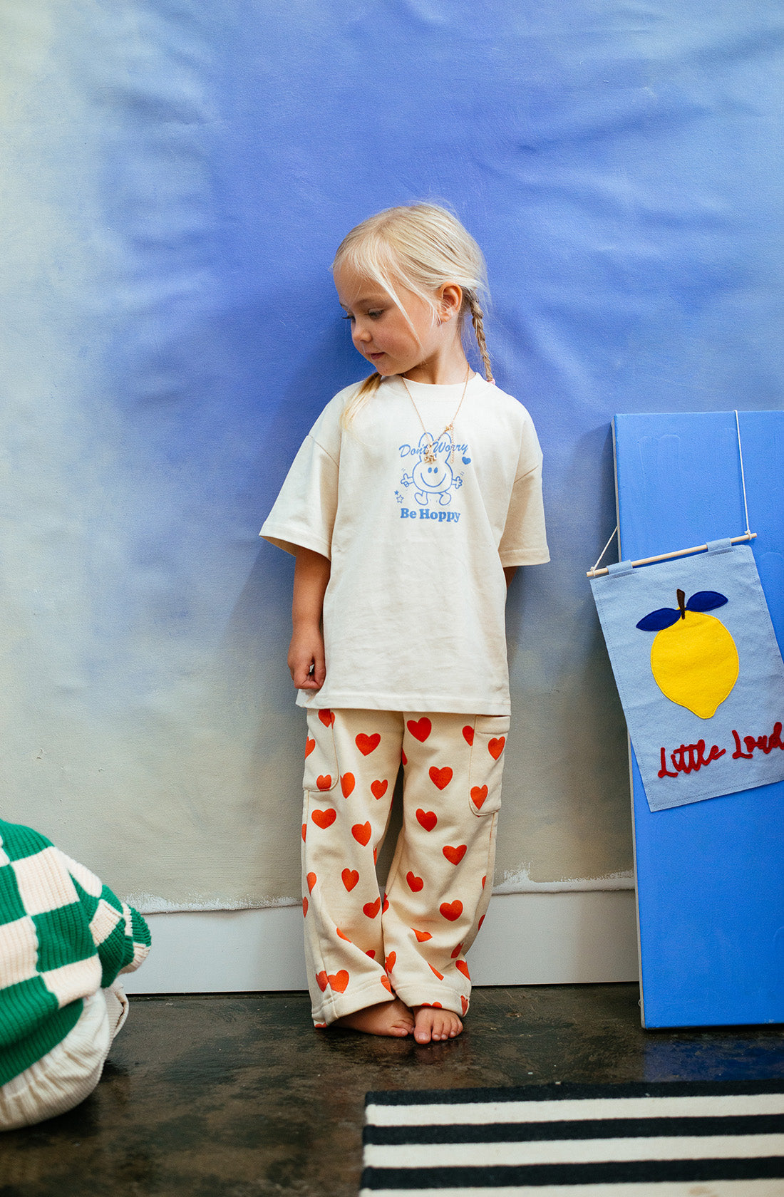 A young child with blonde hair stands barefoot against a blue wall. They wear an oversized white T-shirt with a smiling face and the text "Be Happy," alongside SUNDAY SIBLINGS' ultra-soft, cream-colored Holiday Pants Red Hearts adorned with red hearts. Various colorful objects sit around them.