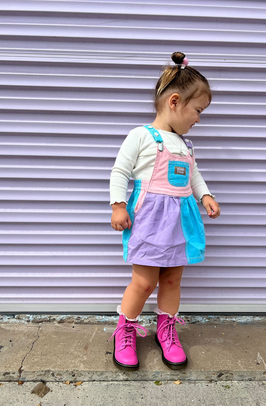 A young child stands in front of a lavender-colored corrugated wall, wearing a white long-sleeve shirt underneath the TWIN COLLECTIVE Bowie Dress Colourblock. This pastel-colored bubble dress features sections in pink, purple, and blue and is crafted from sustainable 100% cotton. The outfit is completed with bright pink boots and a small bun in their hair.