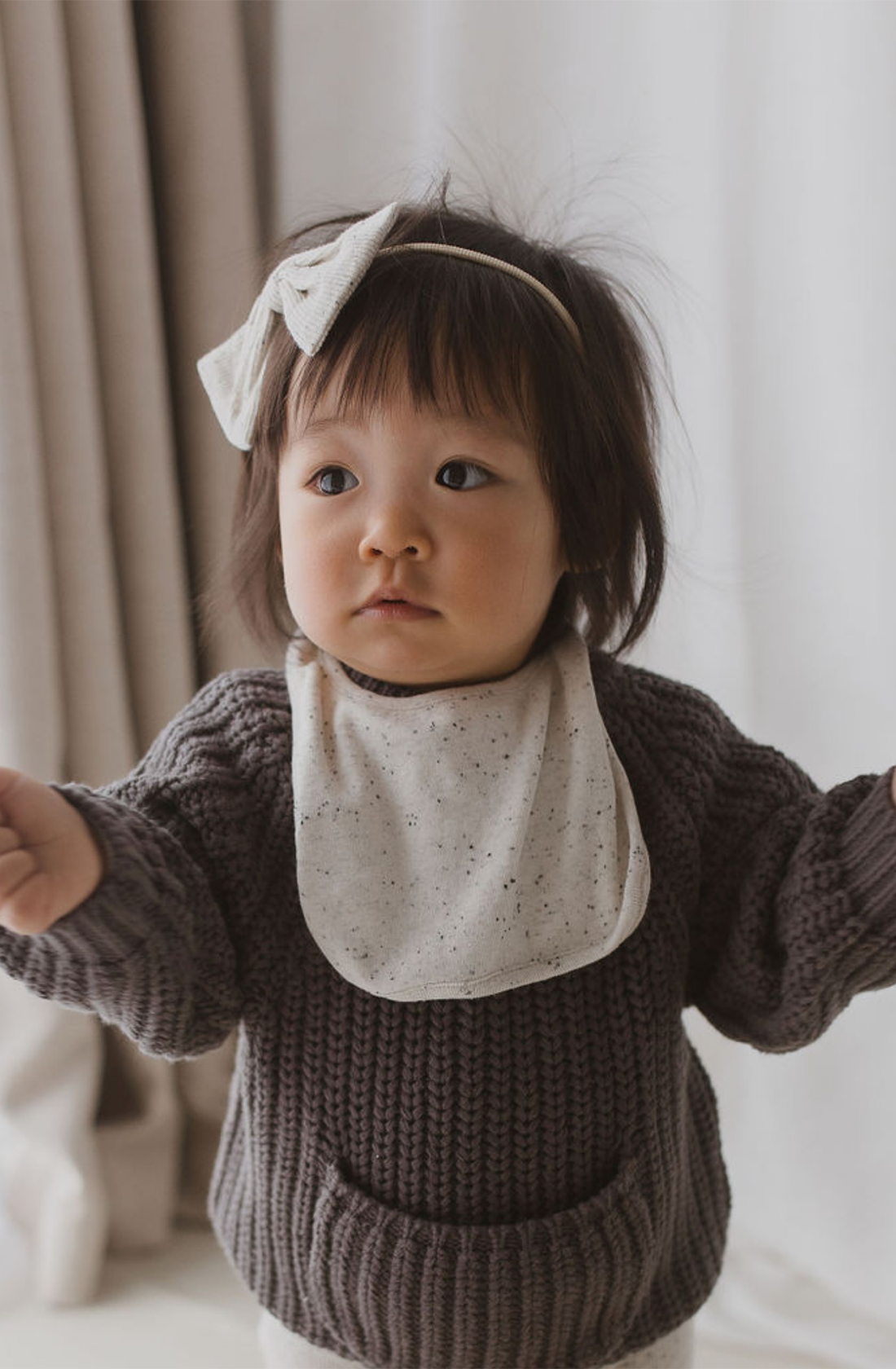 A young dark-haired child, arms outstretched, is dressed in a gray knit sweater and the SUSUKOSHI Boxy Bib Quinoa Speckle made of organic cotton with small speckles. The child wears a headband adorned with a bow and gazes into the distance while light-colored curtains serve as the backdrop.