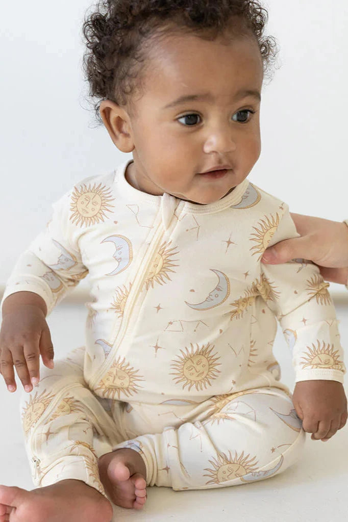 A baby sits on a white surface, dressed in the Celestial Zip Jumpsuit Cream from BAM LOVES BOO, adorned with sun and moon patterns. A hand gently touches the baby's arm, capturing their attention.