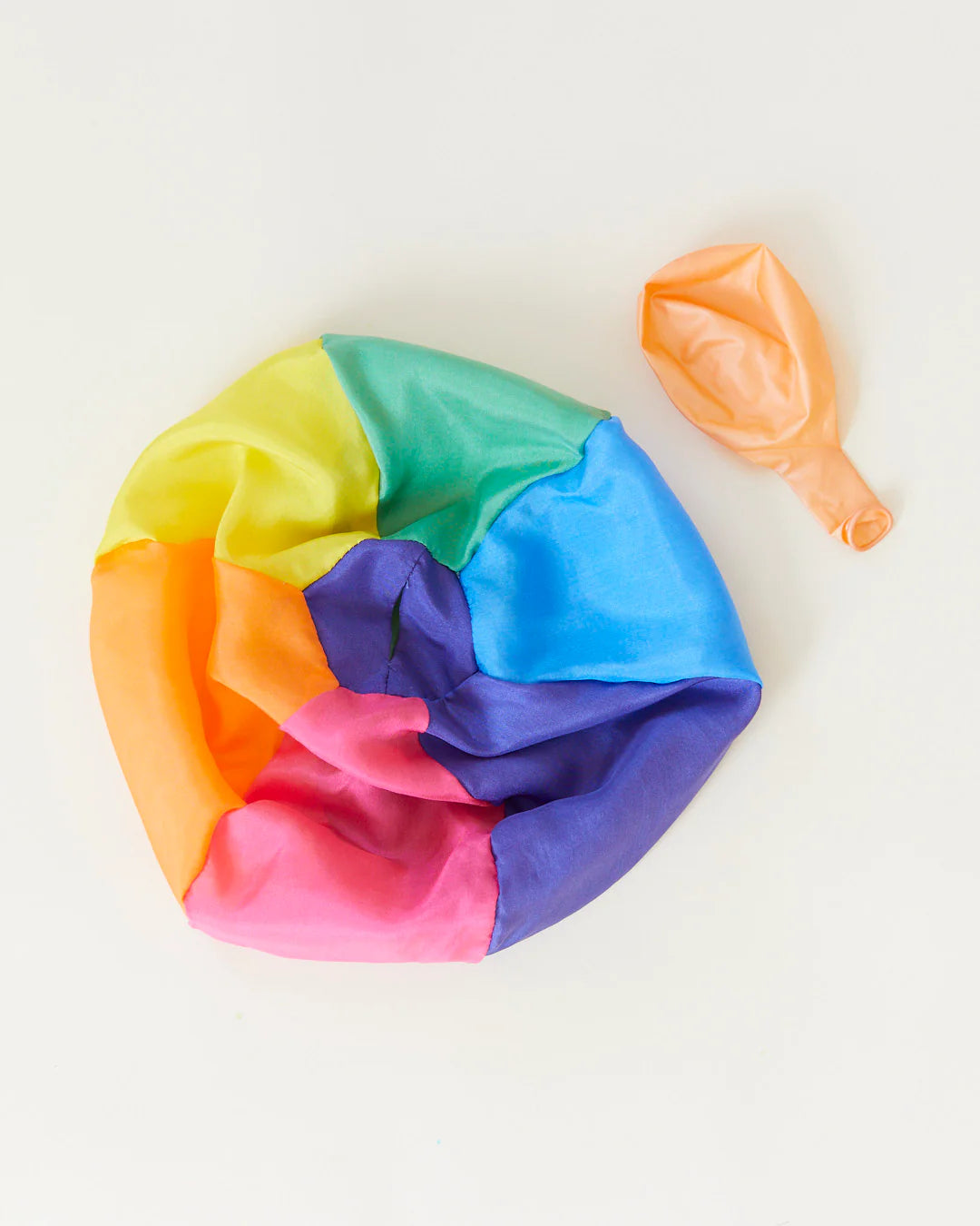 A deflated Rainbow Balloon Ball by SARAH'S SILKS, featuring a multicolored design, and an uninflated orange balloon are placed on a light background, highlighting their potential to transform into vibrant reusable balls for playful adventures.