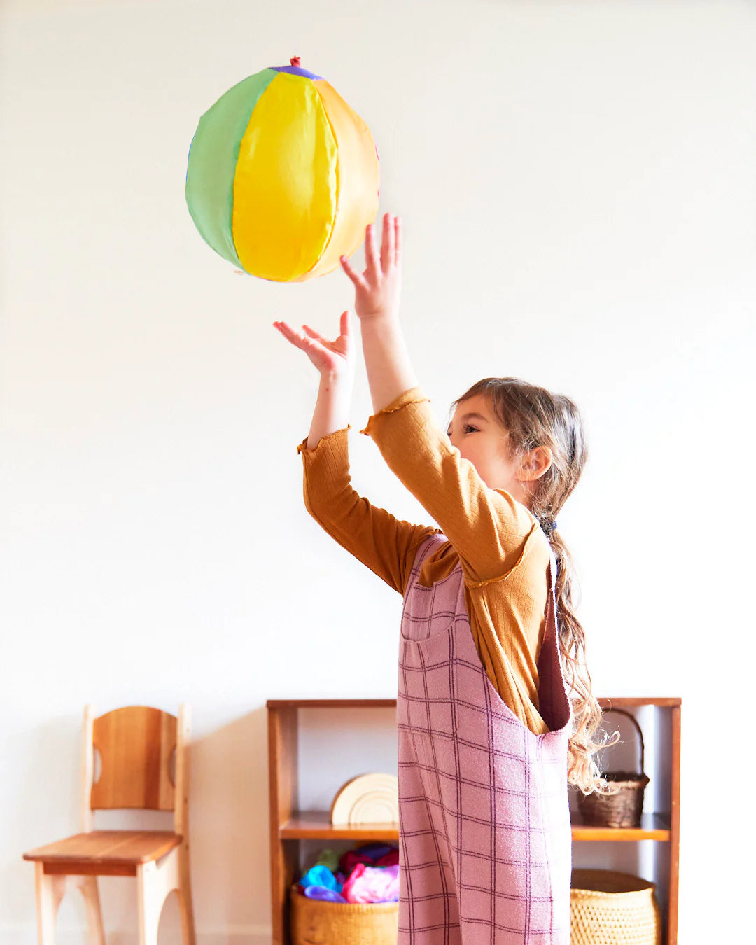 A young girl with long hair and a plaid dress joyfully tosses a colorful Rainbow Balloon Ball from SARAH'S SILKS into the air while playing indoors. Behind her, a wooden shelf with baskets alongside a chair is set against the white wall.