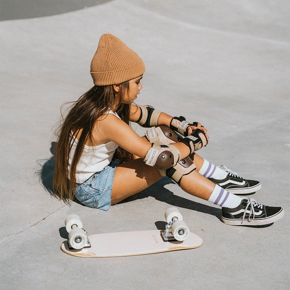 A young girl sitting in the middle of a skate park wearing banwood skateboard protective knee pads, elbow pads and wrist guards in tan/brown.