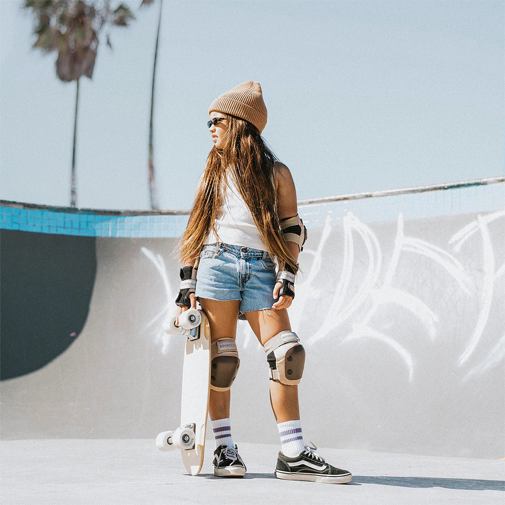 A young girl standing in the middle of a skate park wearing the banwood skateboard protective knee pads, elbow pads and wrist guards while holding her white banwood skateboard.