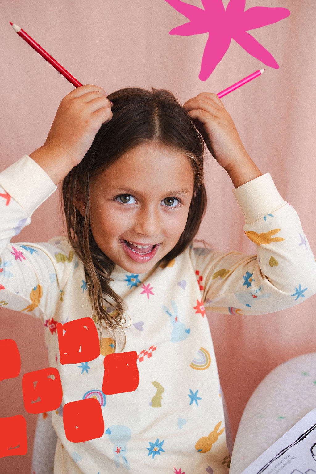 A young girl in a colorful BAY KIDS Kids Sleepwear Set Hoppy Dreams playfully holds red and pink pencils like antennae, smiling at the camera against a vibrant pink background.