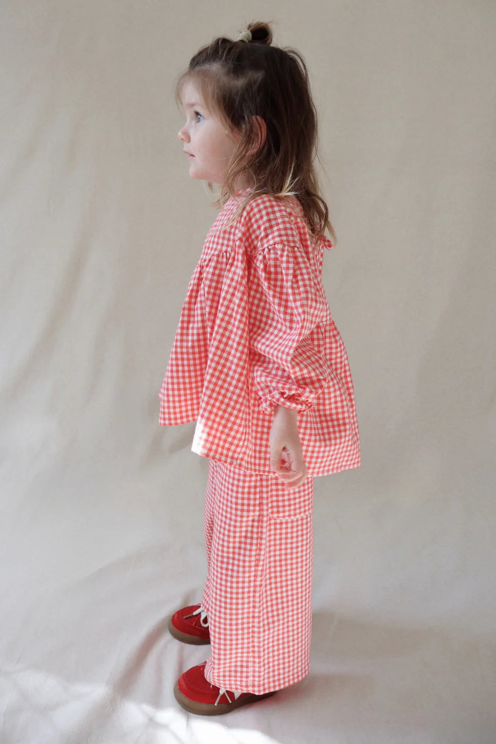A young child stands in profile, wearing the WORN CURATIONS Worn Curations - Gingham Bloom Blouse Chilli, paired with matching pants and red shoes, set against a plain light background.