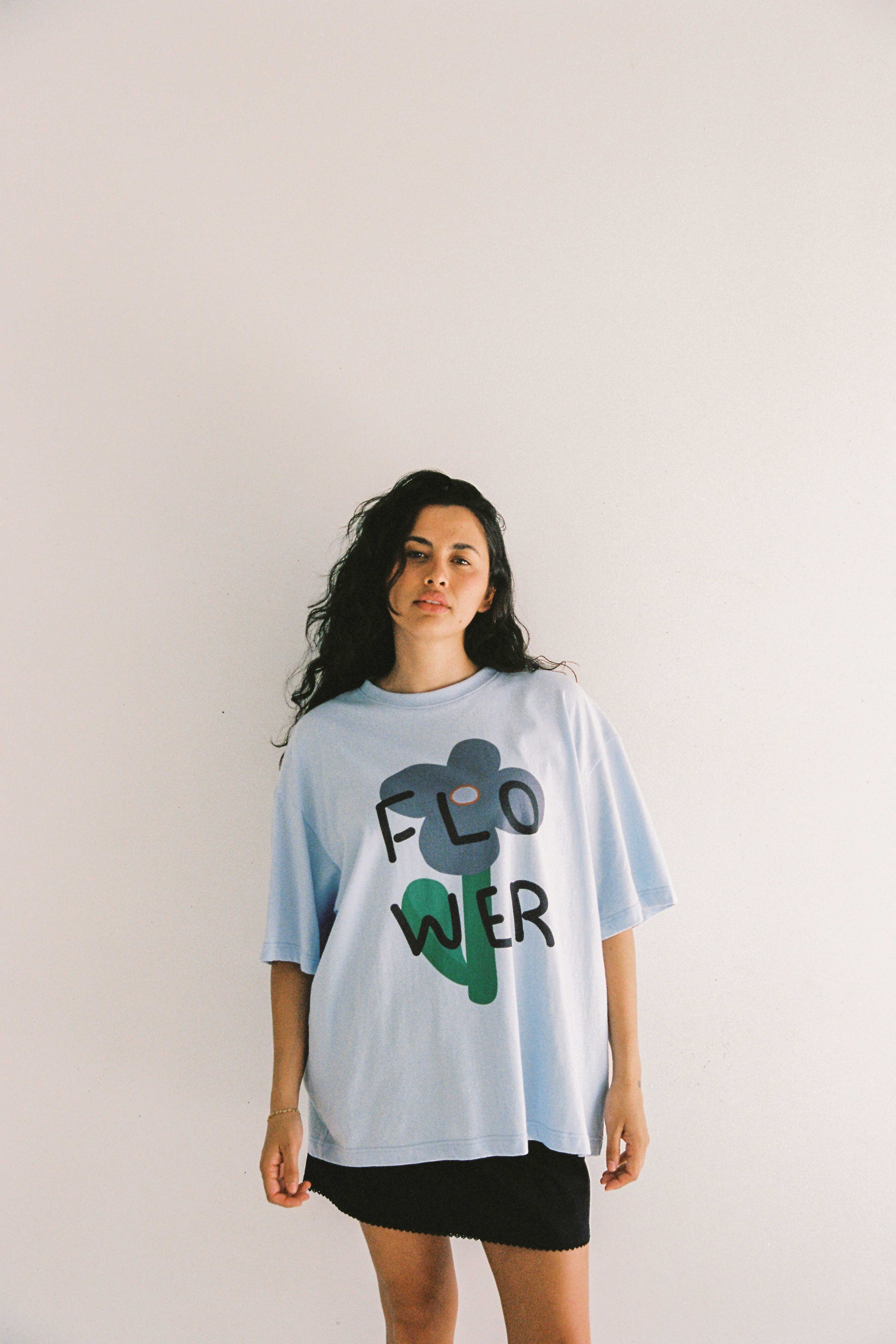 A person with long, wavy hair stands against a plain white background. They are wearing an oversized fit Bloom Tee Light Blue from JUNI, made from organic cotton, featuring the word "FLOWER" printed in bold, abstract letters, and a short black skirt. They look directly at the camera with a neutral expression.