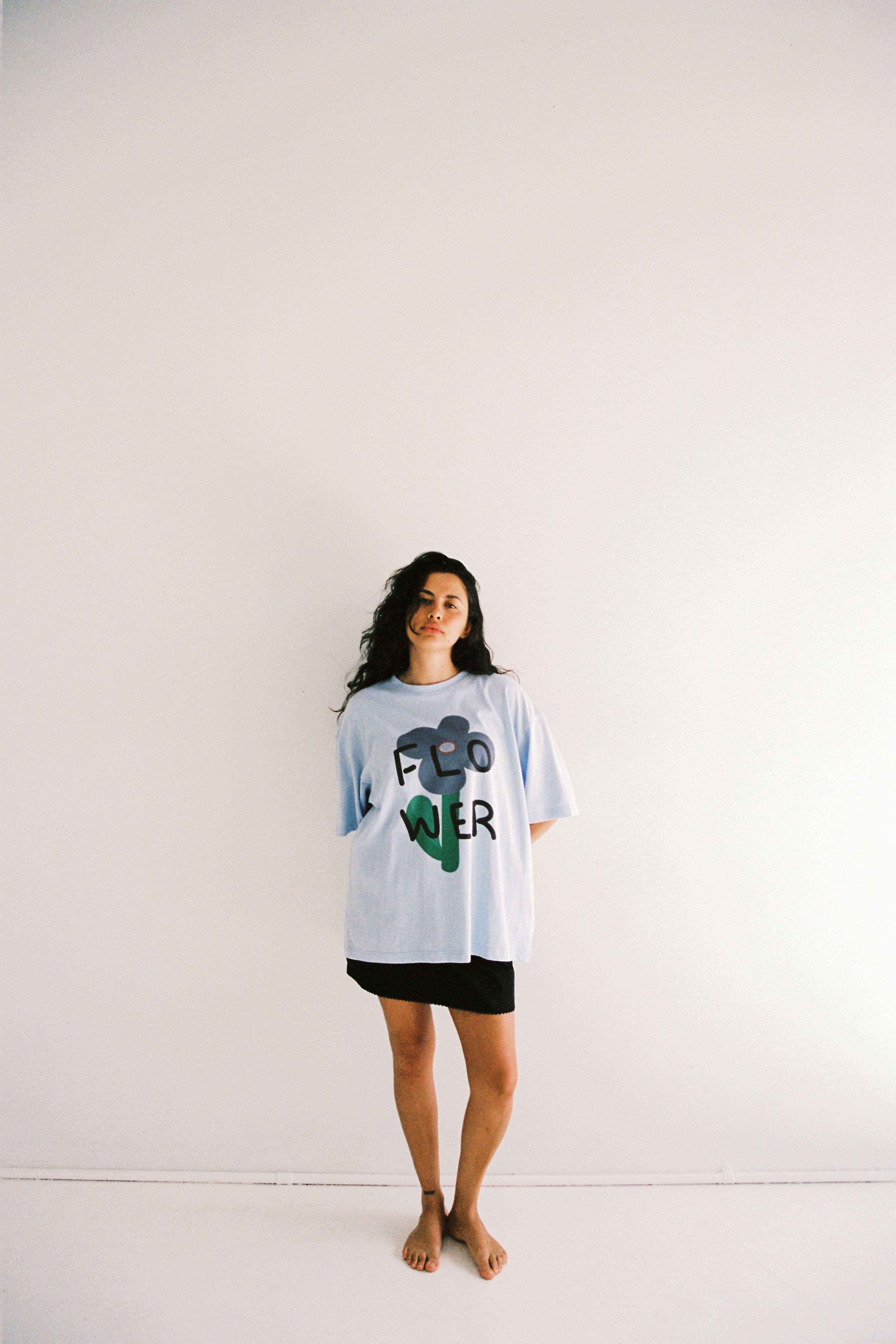 A person with long hair stands barefoot against a plain white wall. They are wearing JUNI's ethically made, oversized fit Bloom Tee in light blue with the word "FLOWER" printed on it in black and green letters, paired with a black skirt. Their hands are positioned behind their back.