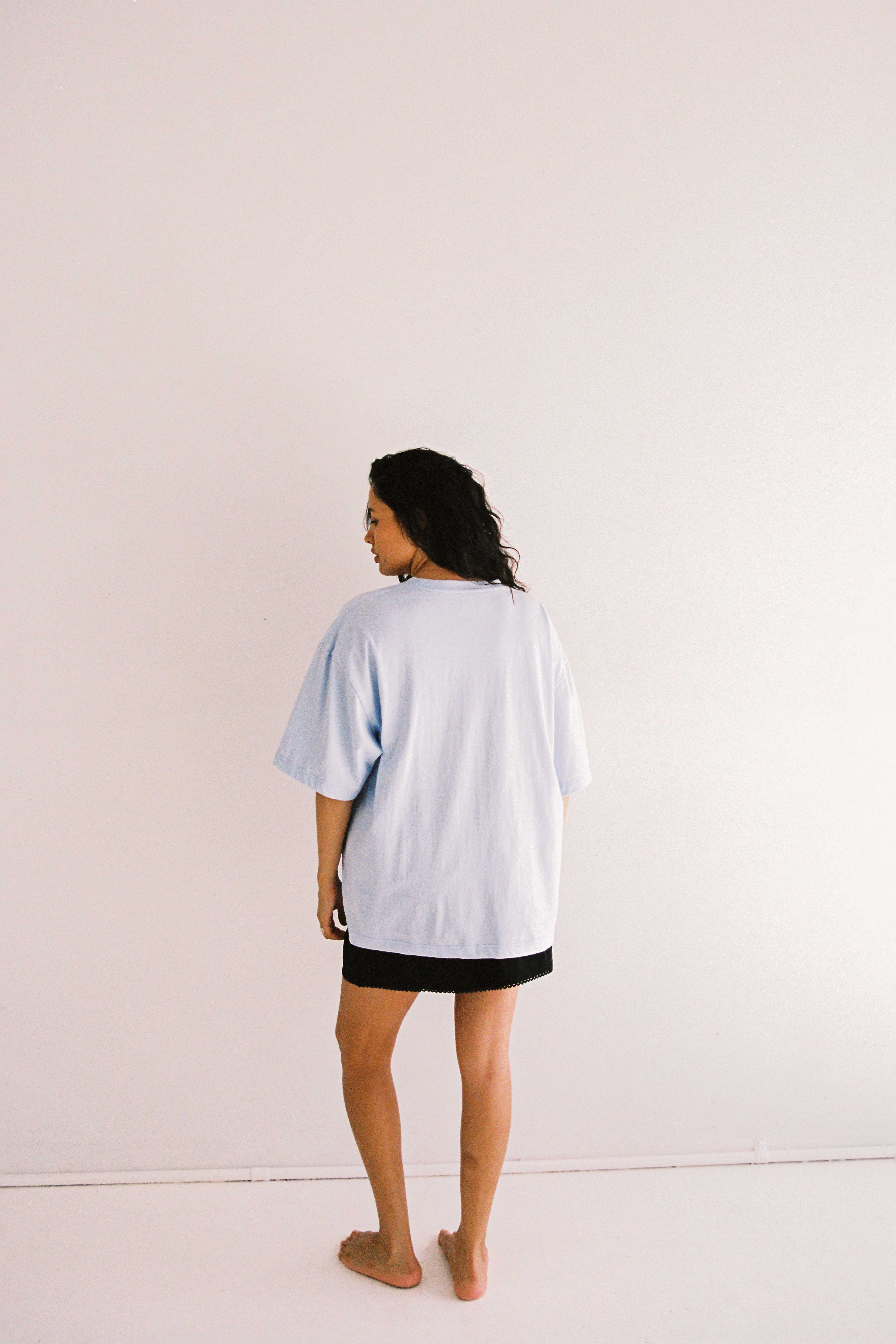 A person with long, dark hair stands barefoot against a plain white background. They are wearing an oversized fit JUNI Bloom Tee Light Blue made of organic cotton and dark shorts. The individual is facing slightly away from the camera, towards their left.