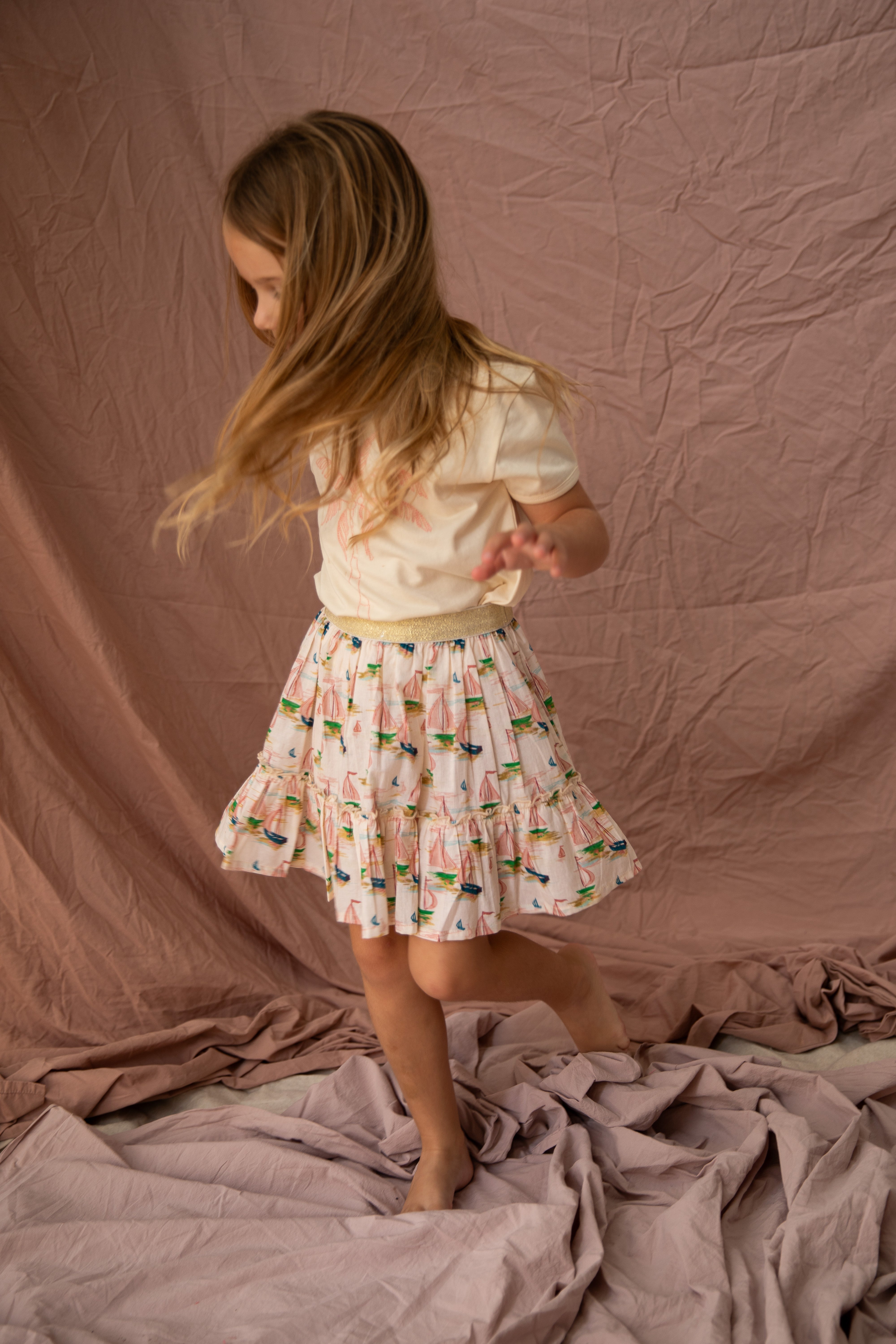 A young girl with long hair wearing a Key Largo Tee in vanilla by BELLA + LACE and a colorful cotton jersey skirt twirls joyfully on a textured pink fabric backdrop, her hair and skirt swirling around her in motion.