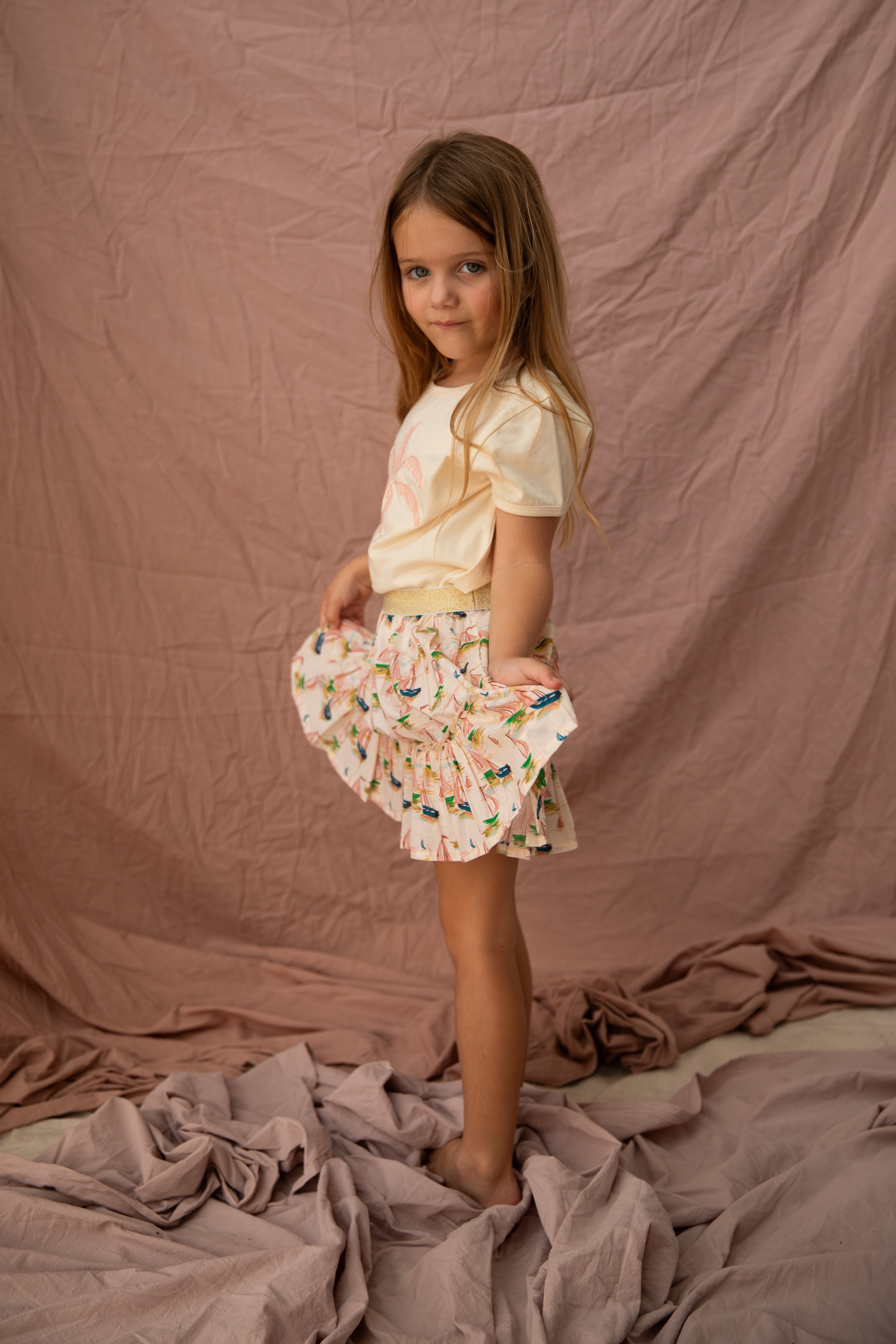 A young girl stands on a crumpled fabric, wearing the Key Largo Tee in Vanilla from BELLA + LACE and a ruffled skirt featuring a colorful pattern. The backdrop is textured mauve, and she holds the skirt slightly out to the side.