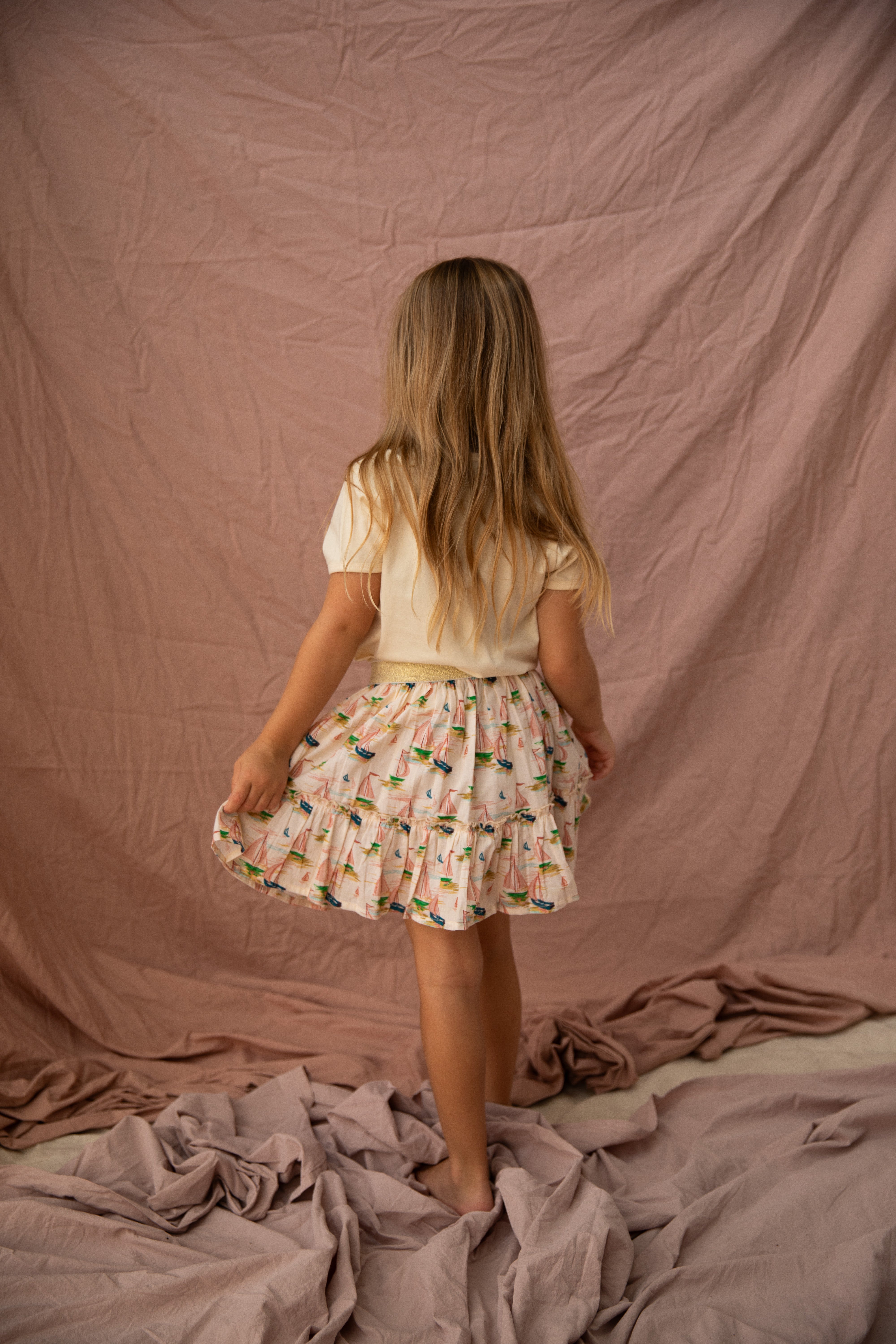 A young girl with long hair stands facing away, dressed in the BELLA + LACE Key Largo Tee Vanilla, a short-sleeved white top paired with a frilly skirt featuring colorful patterns on cotton jersey. She gently holds the edge of her skirt while standing on soft, draped fabric that matches the pink backdrop.