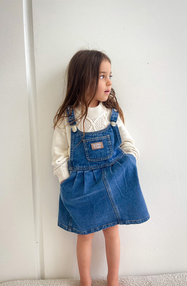 Girl standing against white wall wearing denim dress with knit underneath