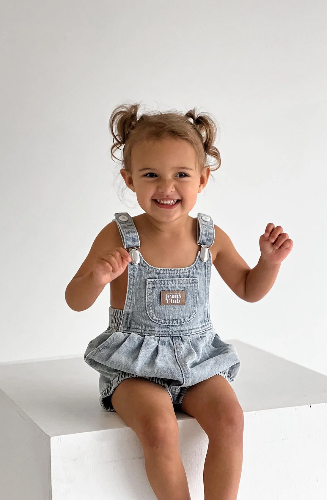 Girl wearing light blue denim romper sitting on white plinth