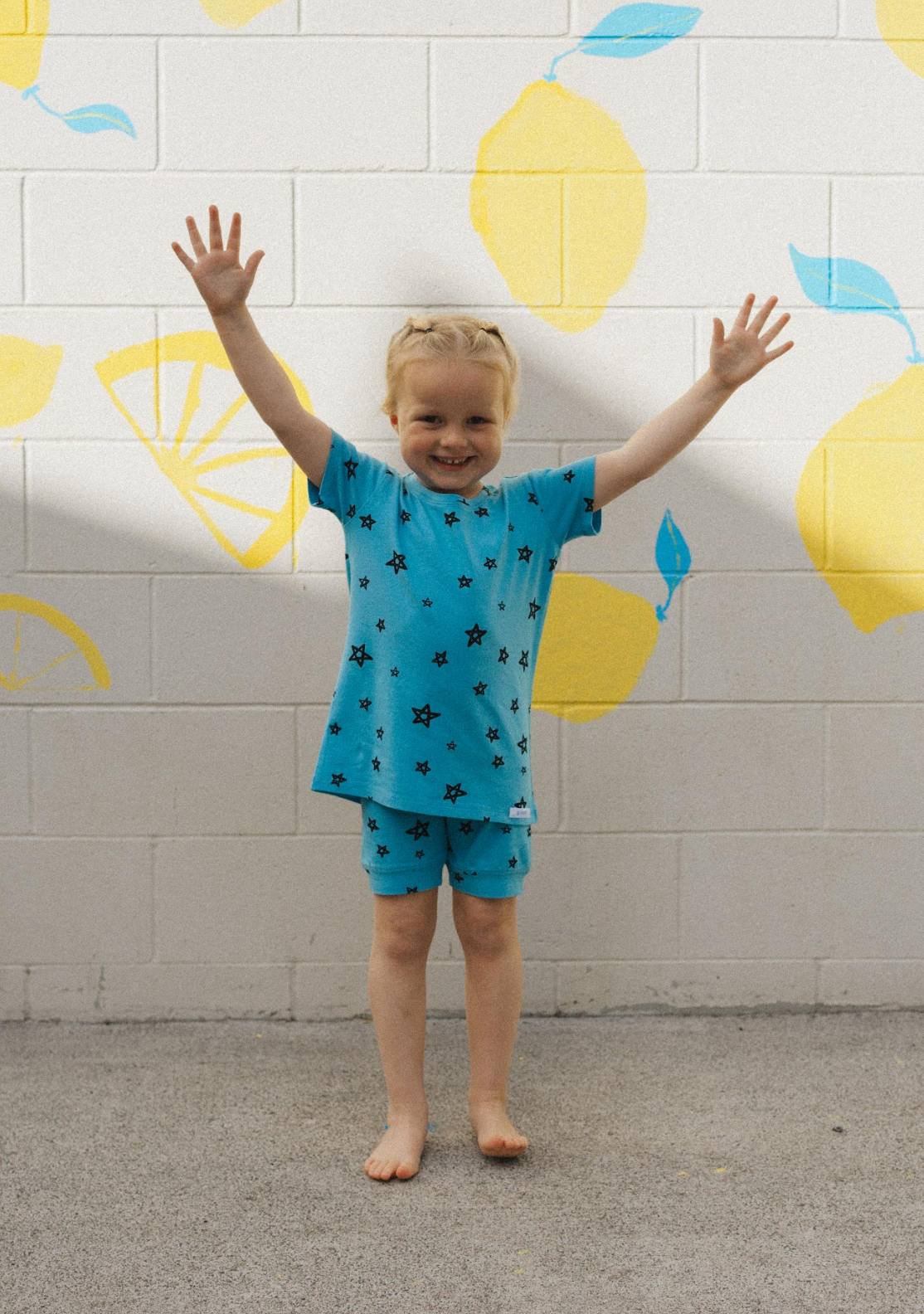 A cheerful child in the Star Shortie PJ Set Breeze by G.NANCY, featuring super soft fabric with star patterns, stands barefoot against a wall covered in yellow lemon designs, joyfully raising both arms.