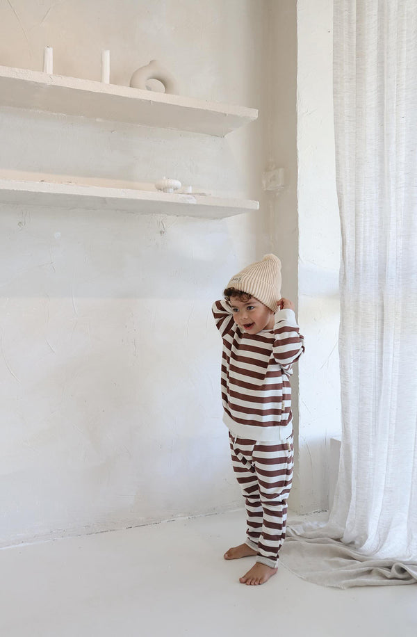 A young boy wearing the Camden chocolate brown striped pullover and matching pants with the cream Bobby G beanie next to see through white drapes and a window.