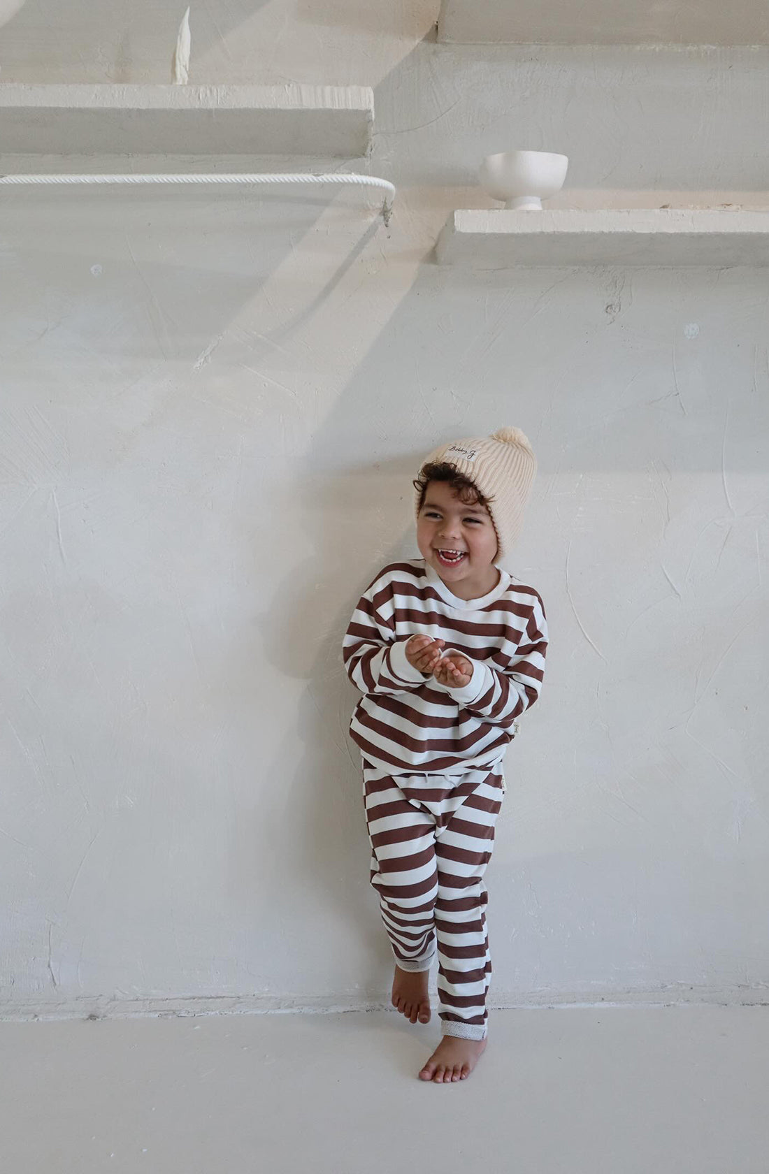 A young toddler wearing the Camden chocolate brown striped pullover and matching pants with the Bobby G cream beanie in an all white room.