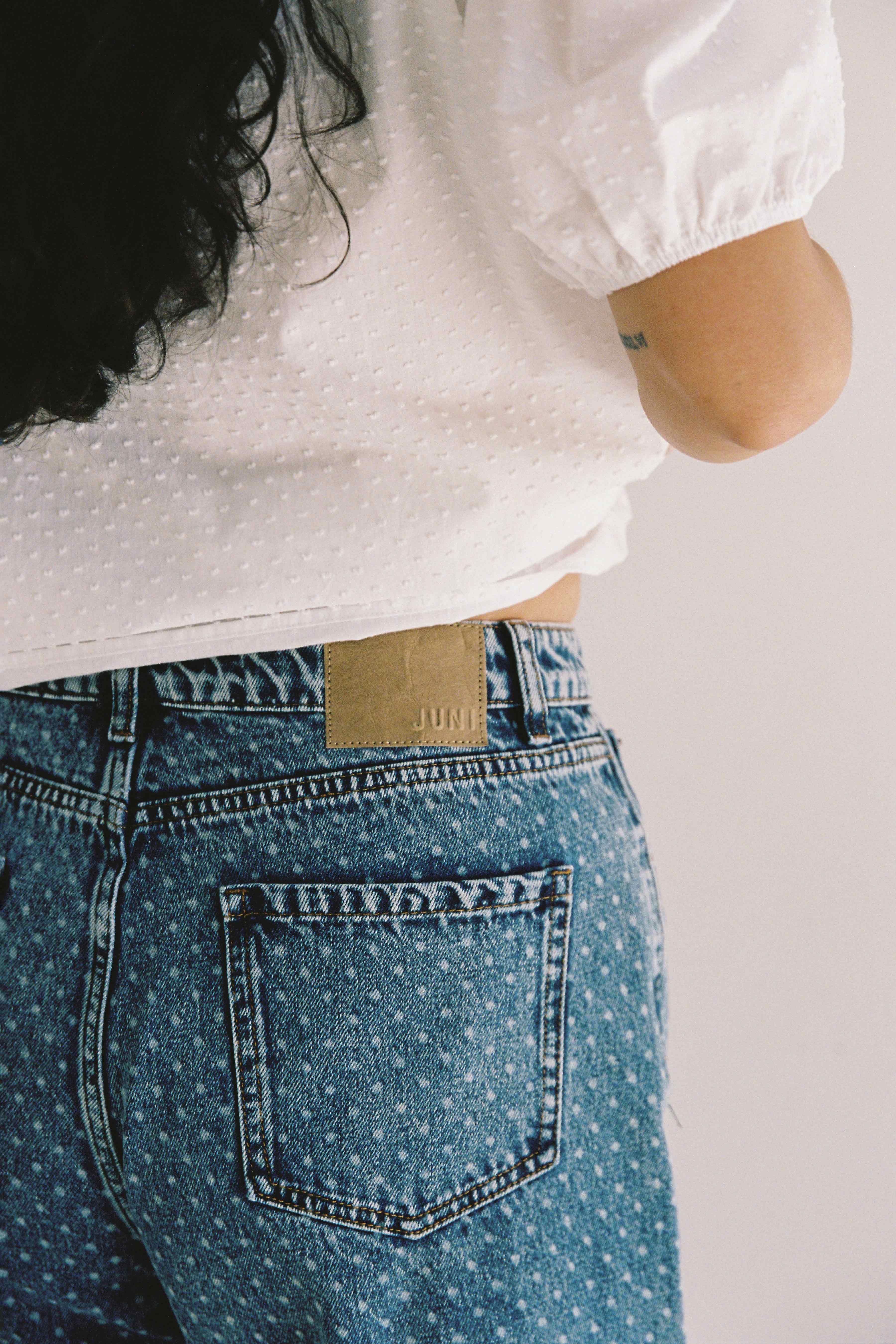 A person with long, dark hair is wearing a white, short-sleeved, textured blouse and a pair of high-waisted Bud Denim Short Spot jeans by JUNI. The blue polka dot jeans are made from cotton denim and feature a leather patch above the back pocket. The image is focused on the back of the person's upper torso and jeans.