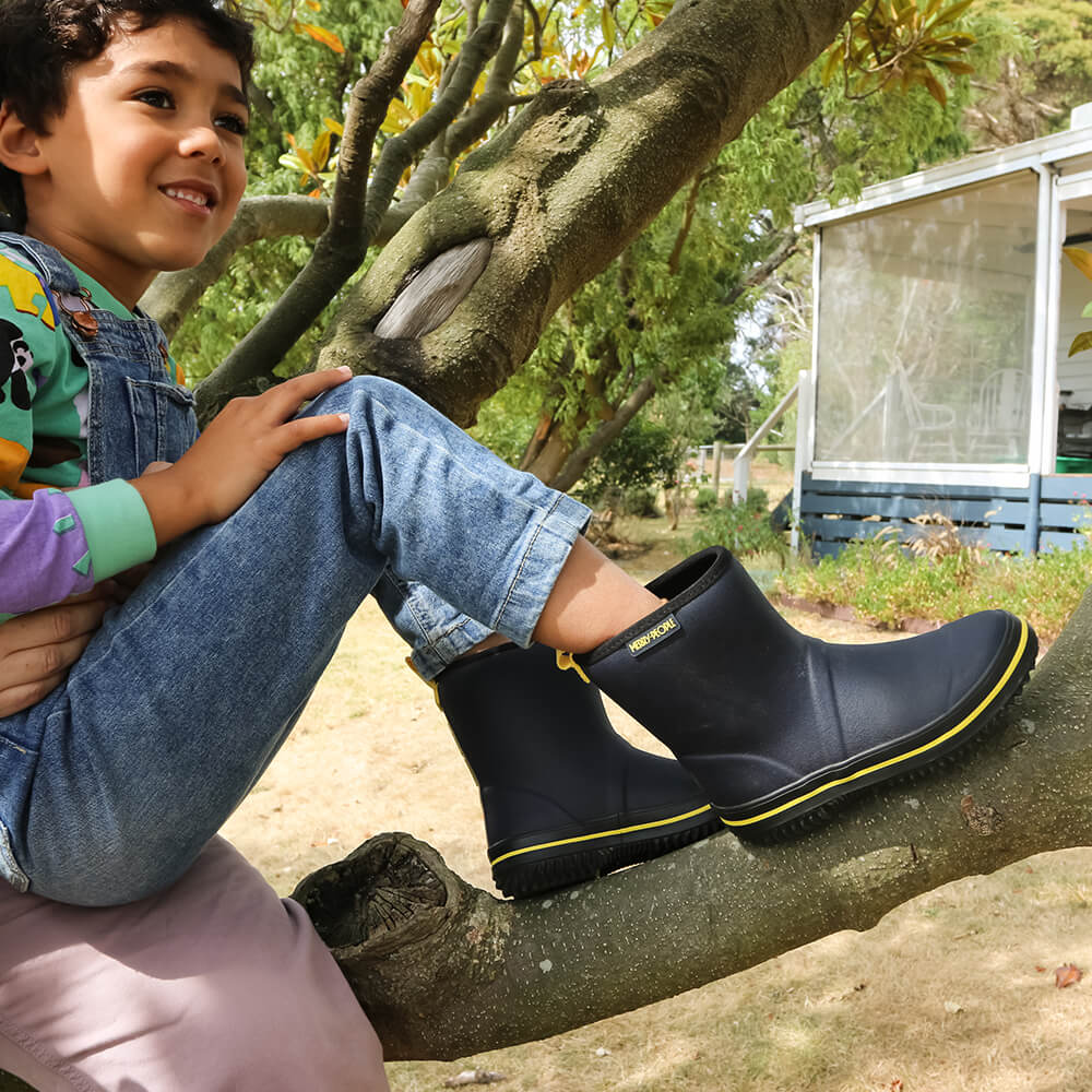 A child wearing Merry People ~ Andie Kids Gumboot Midnight Blue sits on a tree branch. They smile, gazing into the distance, with a vegan-friendly greenhouse nestled among the trees in the background.