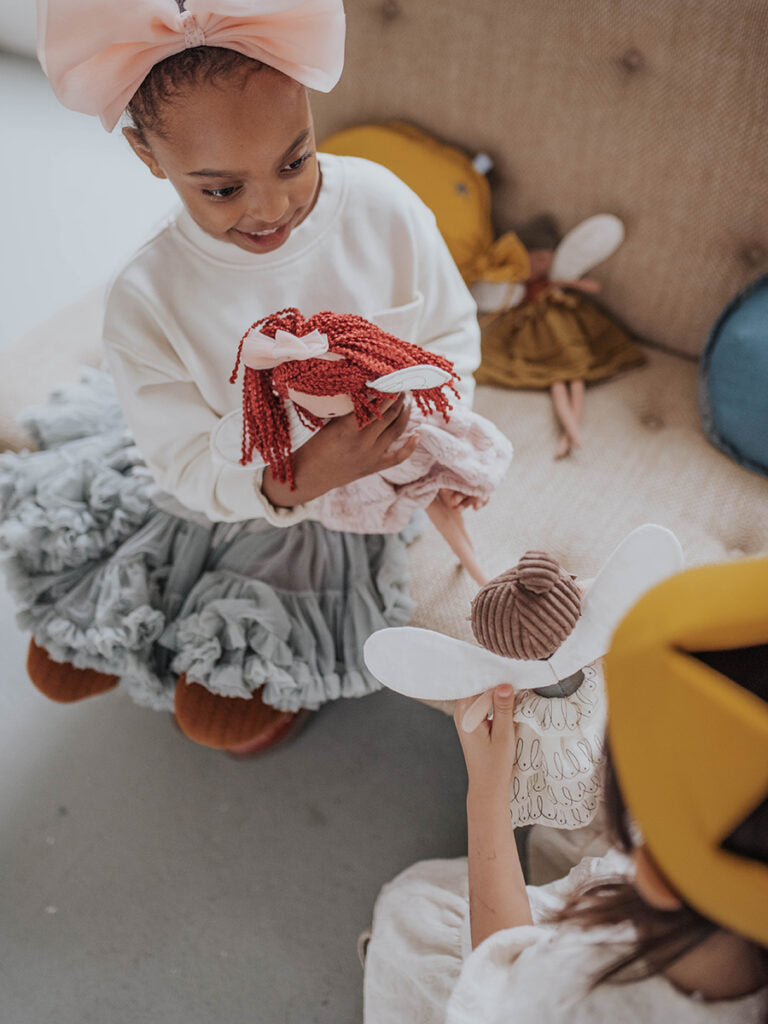 Two children are sitting on a cushioned bench, playing with cloth dolls from Bon Ton Toys. One child with a pink headband and a white top holds PICCA LOULOU's doll with red yarn hair. The other child, partially visible, wears a yellow cap and holds PICCA LOULOU's Fairy Celeste 35cm with white wings.