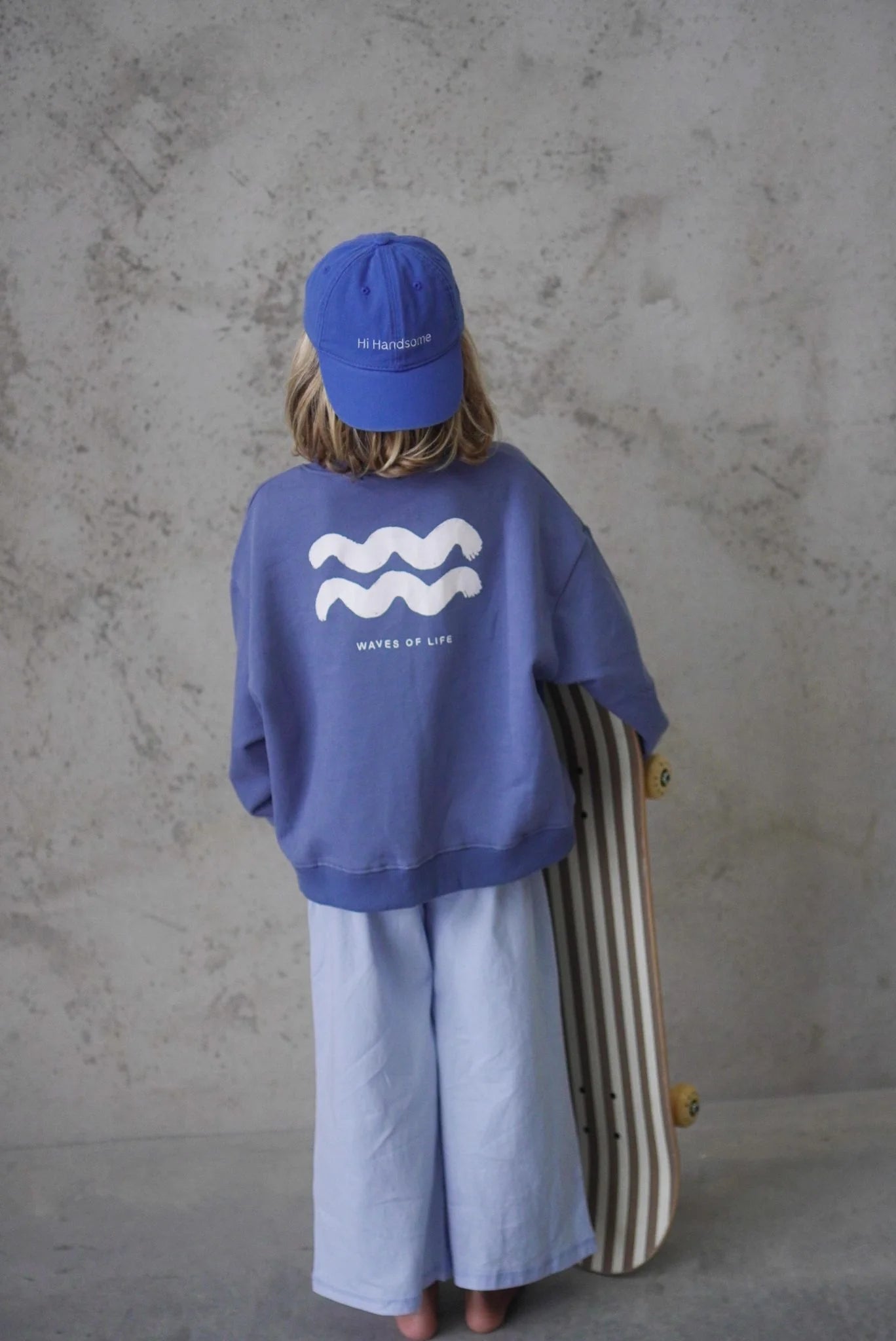 Young boy wearing the relaxed beach pant in blue with a blue cap on backwards and a blue jumper with two white waves on them holding a skateboard.