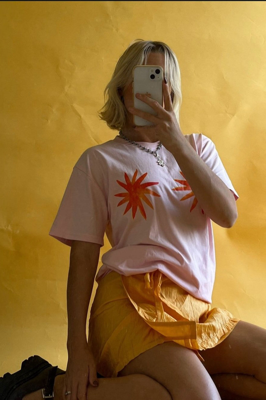 A person with blonde hair takes a mirror selfie against a yellow background. They're wearing the Star Boobies T-Shirt by MAKU FENAROLI, featuring a pink classic fit with red starburst designs that evoke hand-painted artistry, paired with a yellow skirt and accessorized with a necklace.