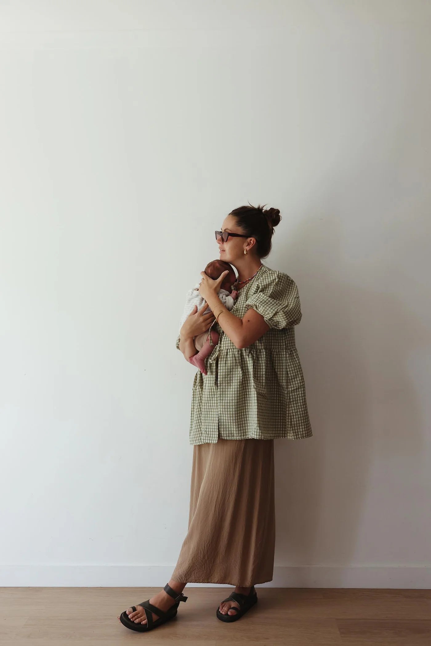 A woman wearing the LITTLE THE LABEL's Mama Cami Blouse in Olive Gingham and a beige skirt stands against a white wall, cradling a small sleeping dog. She accessorizes with sandals and sunglasses, her hair styled in a bun as she gazes to the side.