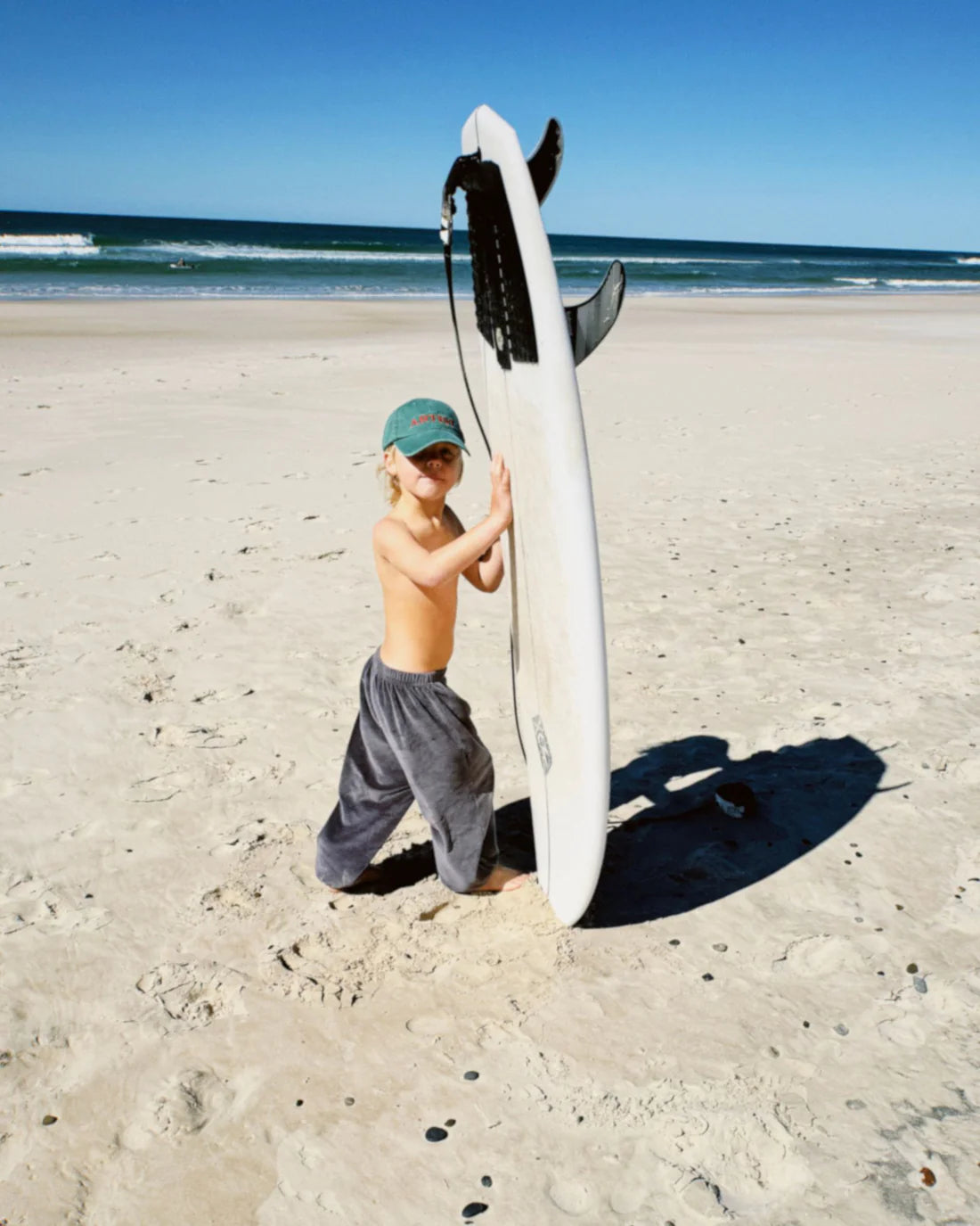 A young child stands barefoot on a sandy beach, holding a surfboard upright. They're wearing gray pants and the Ziggy Zaza ~ Artist Cap Forest Green by ZIGGY ZAZA, with an adjustable brass clasp, against the backdrop of the ocean and blue sky.