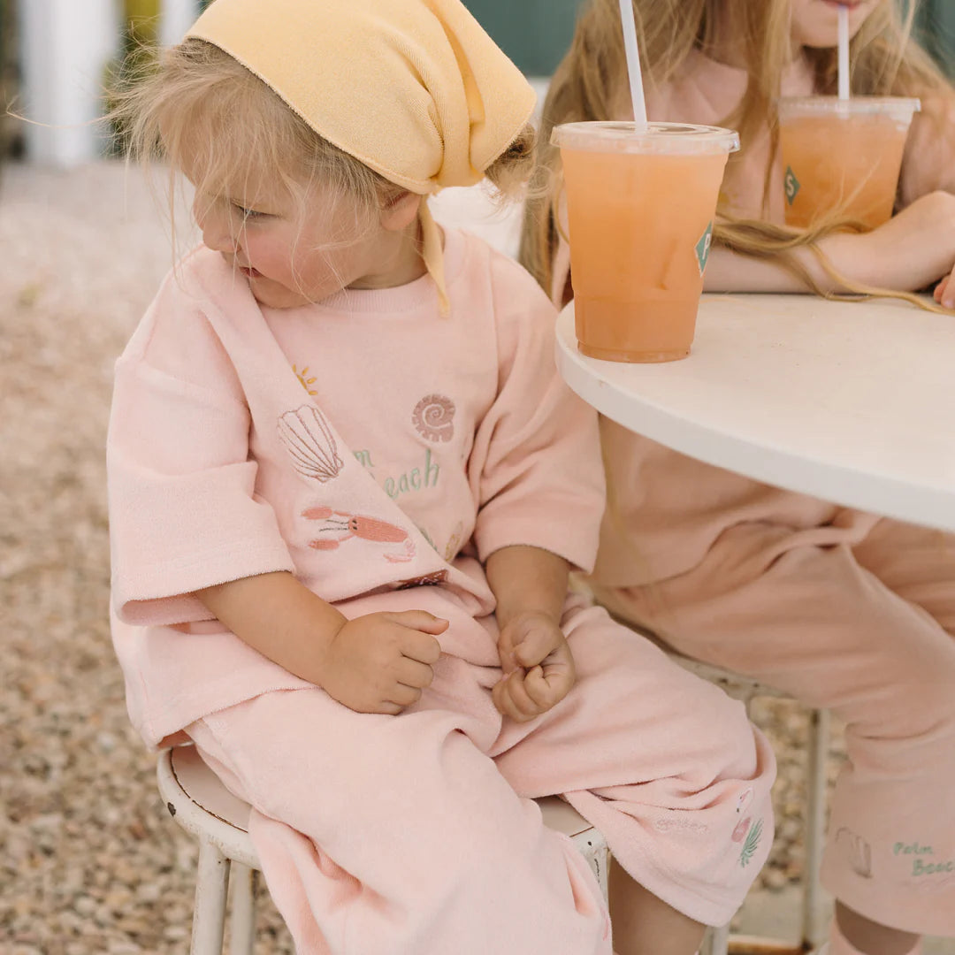 Two children in matching pink outfits sit at a round table outdoors. One wears a yellow headscarf and holds a drink, her Palm Beach Wide Leg Pants by GOLDEN CHILDREN in Flamingo Pink adding to their charm. The table has two plastic cups with straws filled with an orange beverage, while the ground is covered in small pebbles.