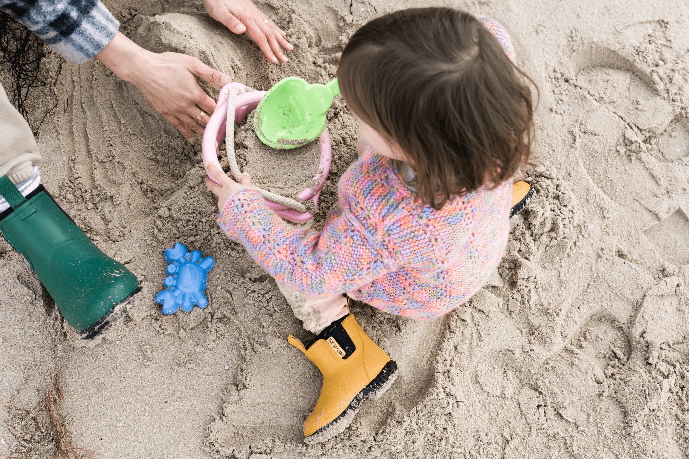 A child in a colorful sweater and Merry People ~ Bobbi Kids Gumboot Mustard Yellow plays with pink and green beach toys on the sand, assisted by an adult's hands. A blue crab-shaped mold is nearby.