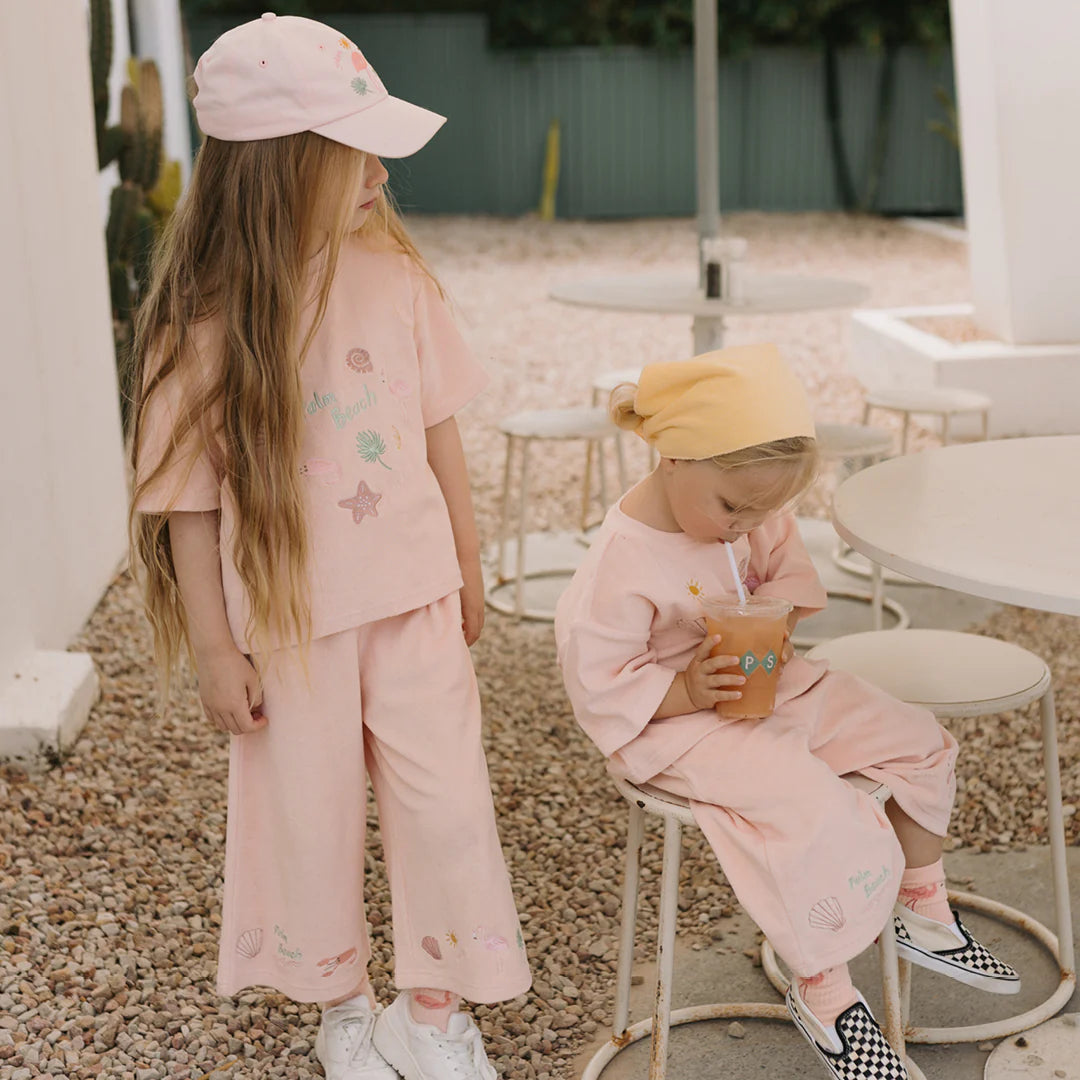 Two young children in matching pastel outfits from the GOLDEN CHILDREN brand, with the Palm Beach Mid Sleeve Tee Terry Towel in Flamingo Pink adding a touch of charm. The older child, sporting a cap, stands next to a seated toddler wearing a yellow headscarf. They are on a patio with white chairs, and one child is holding a cup, highlighting the tee's delicate embroidery details.