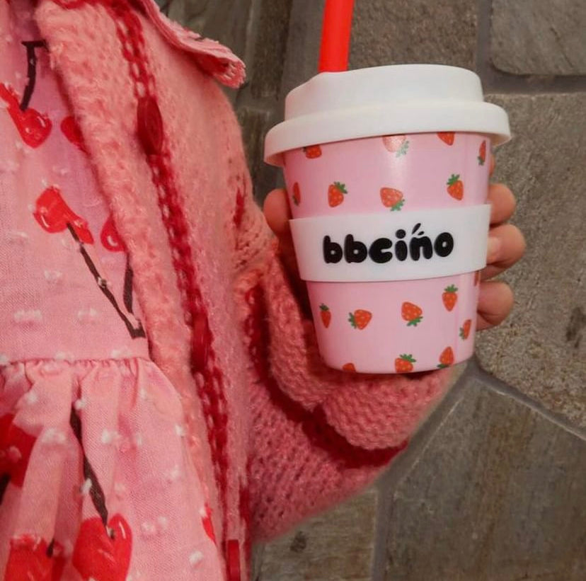 A little girl holding a BBCINO reusable babycino 120ml cup in pink with strawberries scattered over the cup. It has a red straw and a white lid with a white band around the cup with the black BBcino logo on it.