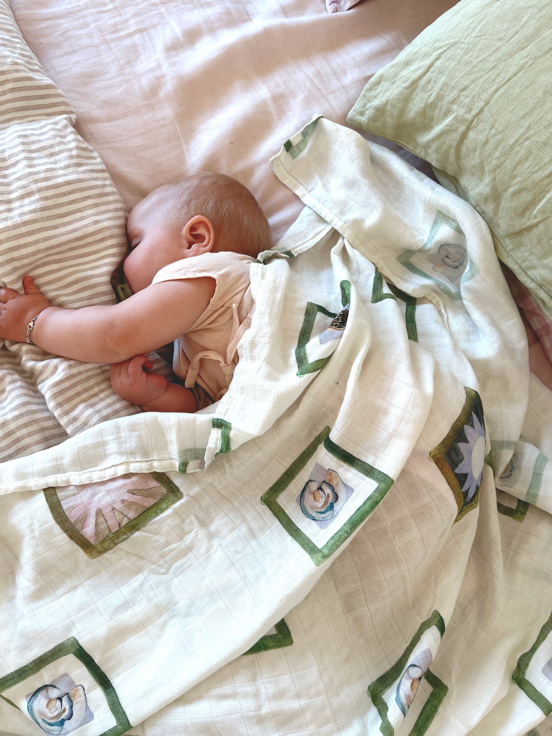 A baby sleeps peacefully on a bed, partially covered with the Annie Wrap from MILKY DESIGNS' floral collection. The baby rests on a striped pillow, with a light green pillow nearby.