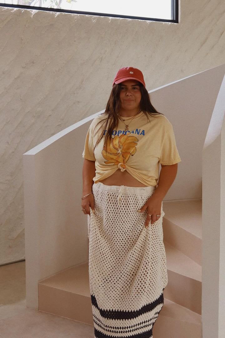 A person wearing a yellow "Tropicana Banana" unisex tee by MAKU FENAROLI and a red cap stands on a staircase. The 100% cotton shirt complements their knitted long skirt. The background features white walls and a large window.