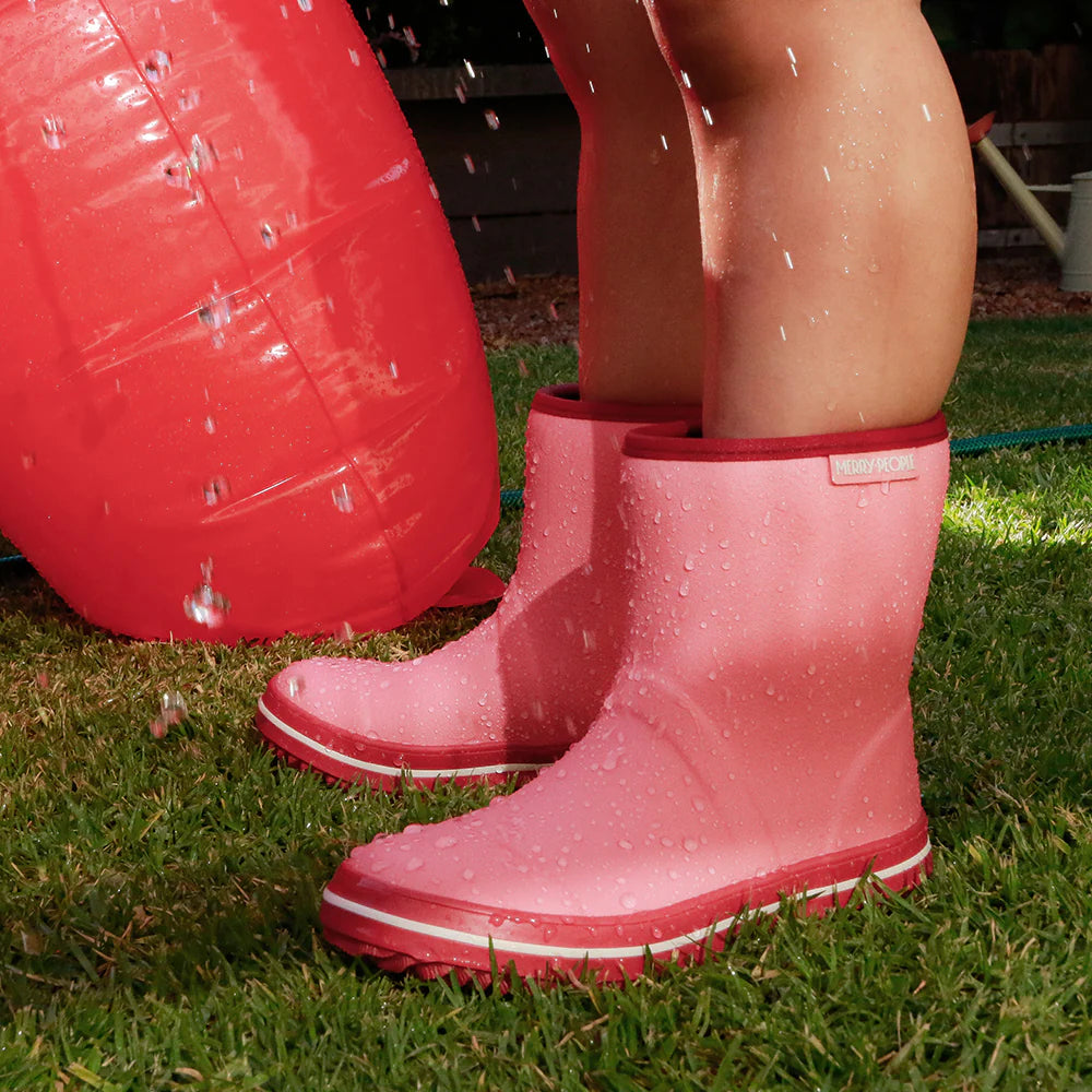 A child in Merry People ~ Andie Kids Gumboot Bubblegum enjoys water splashing on grass, while a vibrant red inflatable ball enhances the fun. Ideal for adventurous little ones who love rainy day play!.