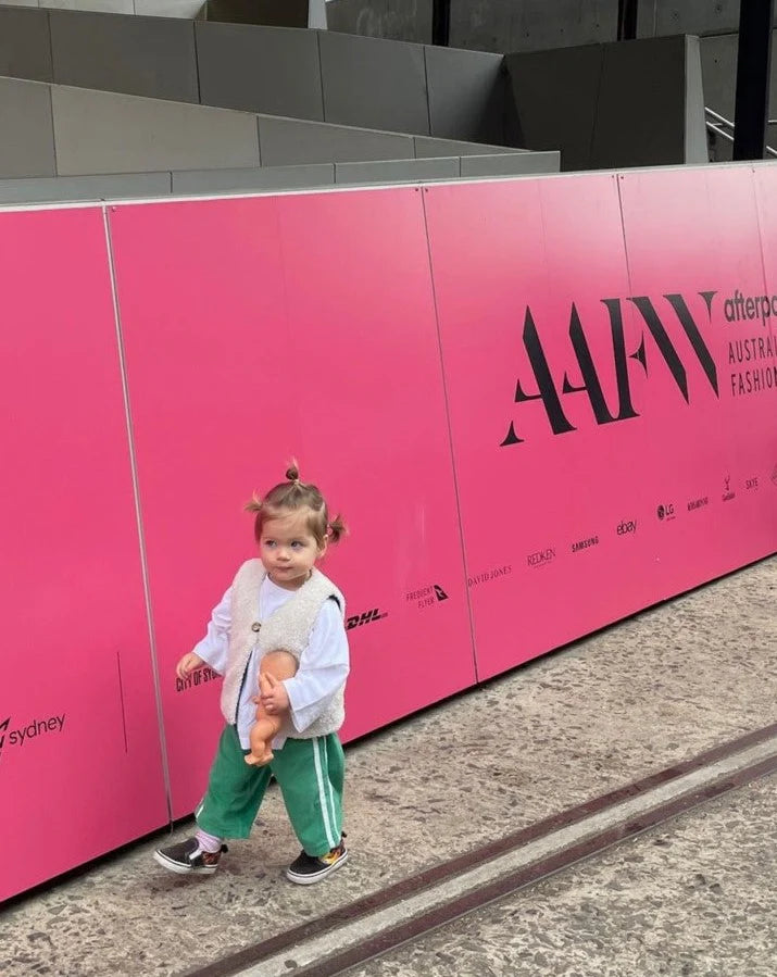 A child in pink hair clips and a white jacket holds a doll near a bright "AAFW" billboard, likely at a fashion event, showcasing style with ZIGGY ZAZA's Ziggy Zaza ~ Twin Stripe Terry Pant Verde. The ground is concrete with barriers visible in the background.