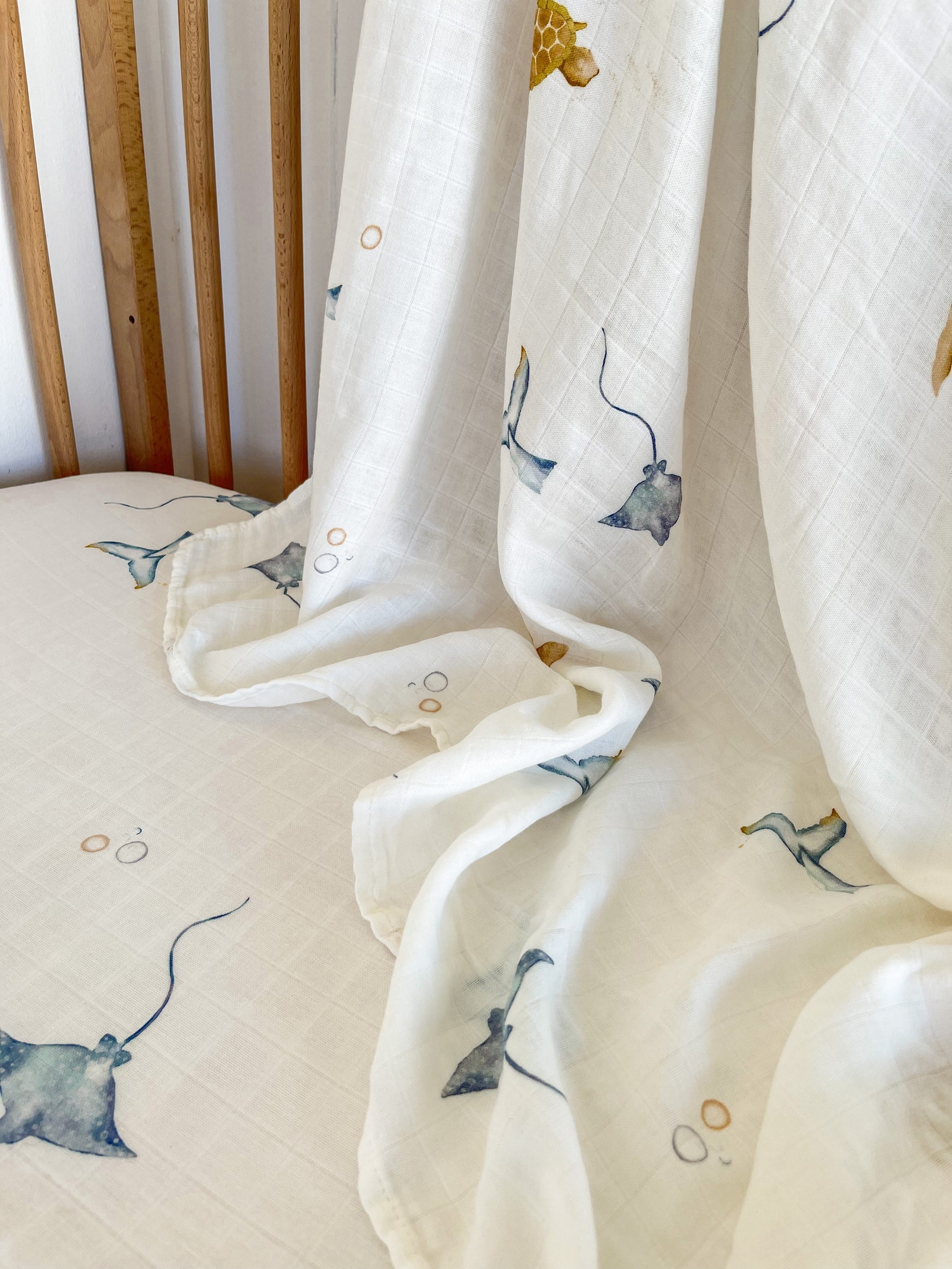 MILKY DESIGNS' Sea Creatures Wrap features a white fabric with exceptional breathability, draped over a wooden chair. It has a subtle grid pattern adorned with stingrays, fish, and bubbles in soft blue and mustard hues for a serene oceanic theme.