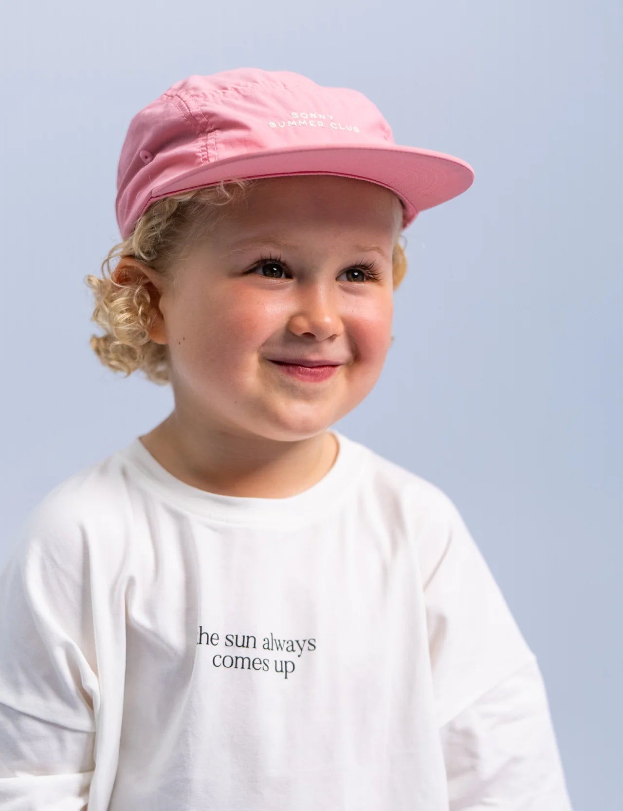 A cheerful child with curly hair is smiling, wearing the Summer Club Cap: Flamingo by SONNY LABEL, a pink cap featuring an adjustable velcro strap, and a white T-shirt that states "the sun always comes up." The background is a soft blue.