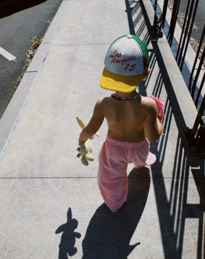 A child wearing Ziggy Zaza's Twin Stripe Terry Pant in pink strolls on the sidewalk with a green and yellow cap, holding a stuffed toy. The shirtless figure casts a shadow, sunlight outlining the toy's shape on the pavement.